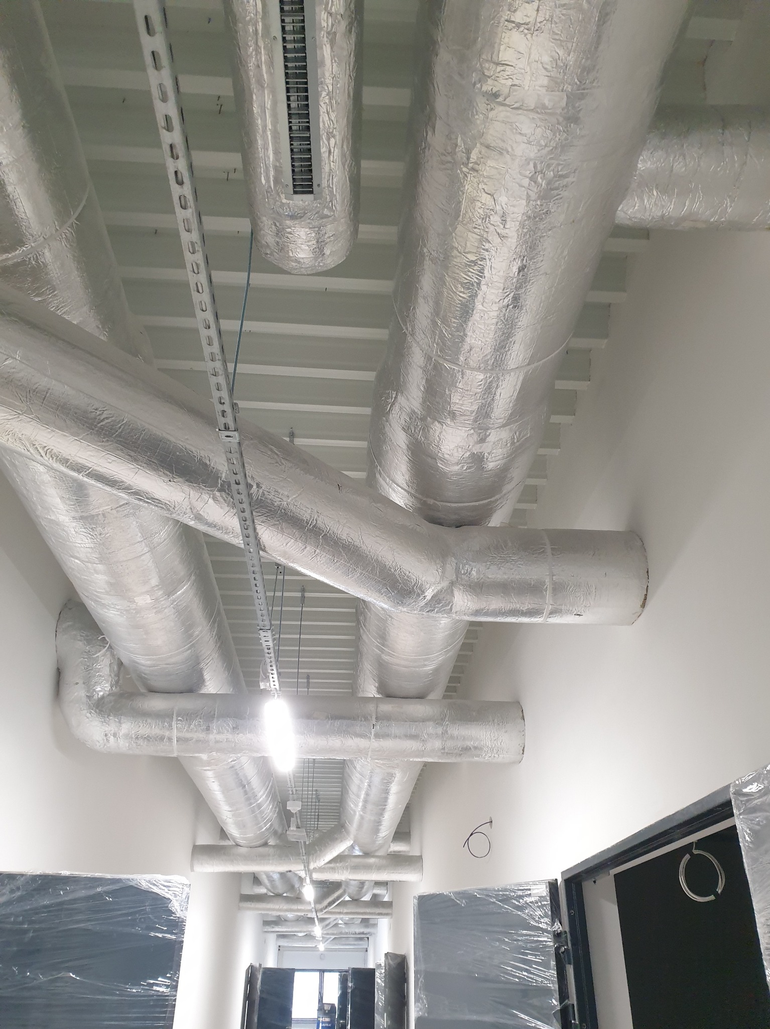 Srebrne, zaizolowane przewody wentylacyjne pod sufitem w jasnym pomieszczeniu. Widoczne mocowania i elementy konstrukcyjne sufitu. Na dole drzwi w folii ochronnej.