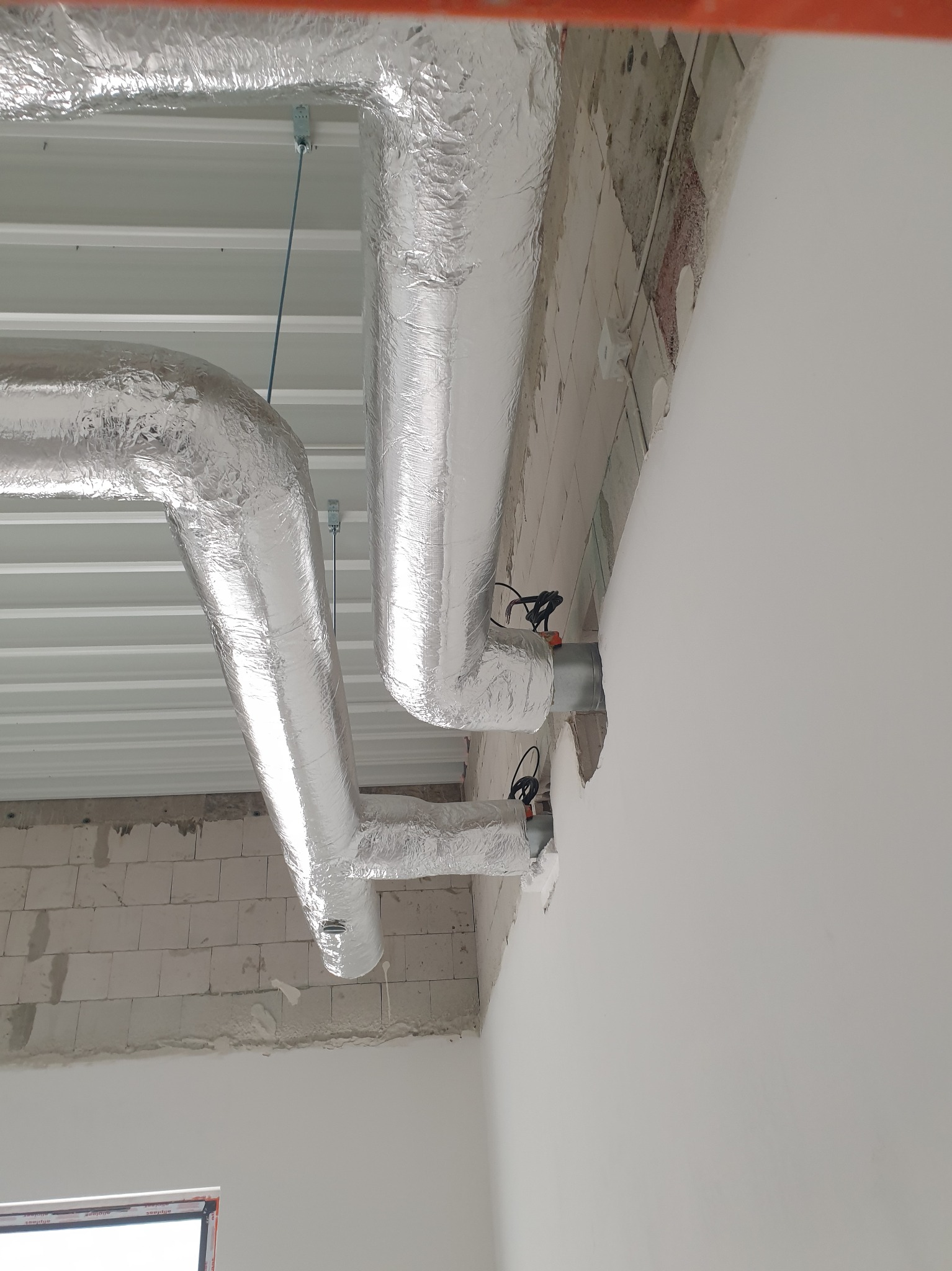 Dwie rury wentylacyjne w srebrnej izolacji, zamontowane pod białym sufitem w surowym wnętrzu. Widoczne przewody i fragment ściany z białego pustaka.