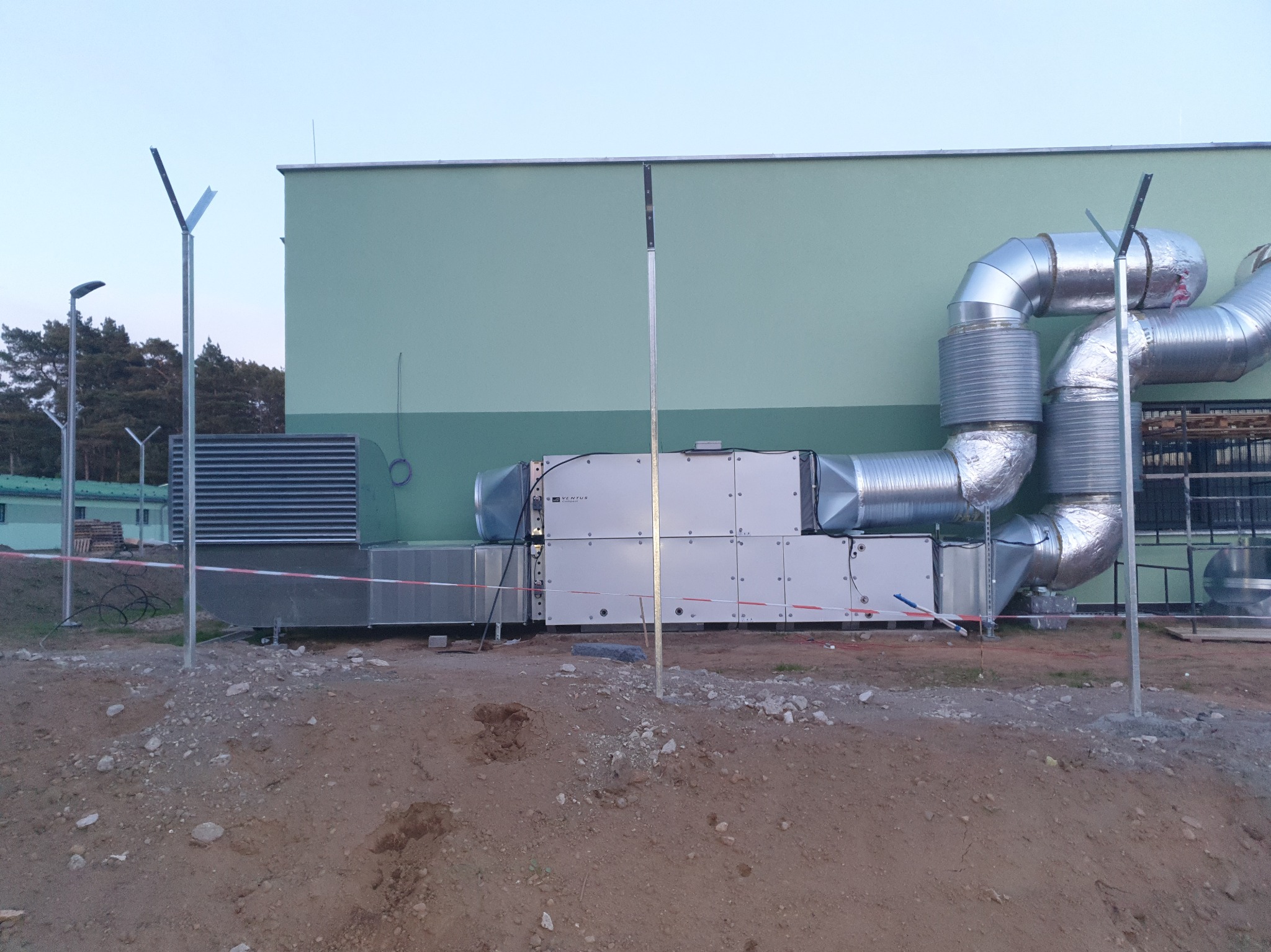 Kompletny system wentylacyjny z srebrnymi rurami na zewnątrz budynku, zabezpieczony taśmą ostrzegawczą. Widoczne słupy oświetleniowe i drzewa w tle.