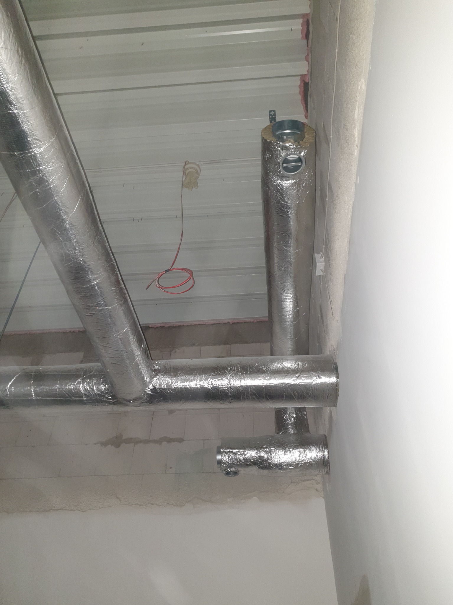 Izolowane rury wentylacyjne pod sufitem z blachy trapezowej. Widoczne przewody elektryczne i fragment ściany z niedokończonym tynkiem.