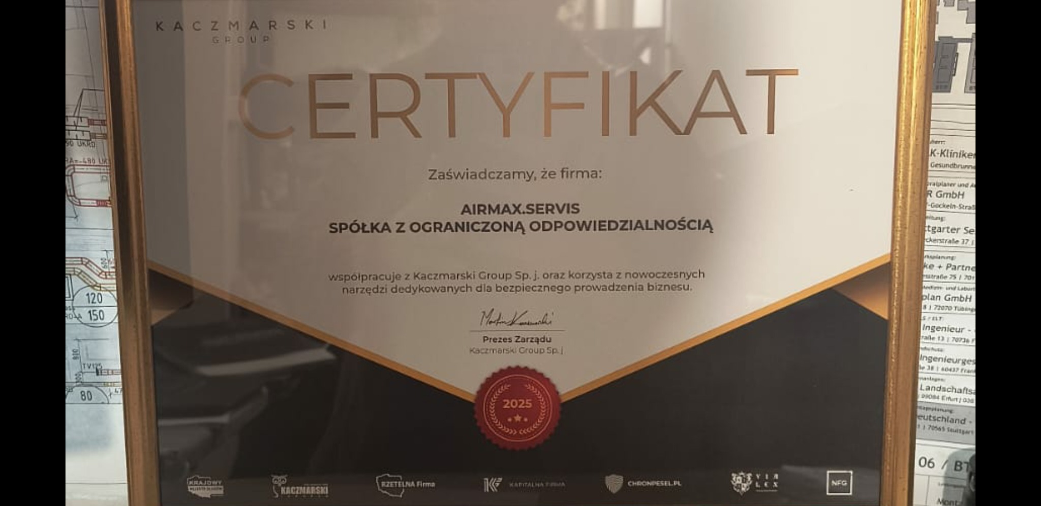 Certyfikat firmy AIRMAX.SERVIS Sp. z o.o. o współpracy z Kaczmarski Group, w złotej ramce, leżący na tle planu technicznego. Sygnatura 2025.