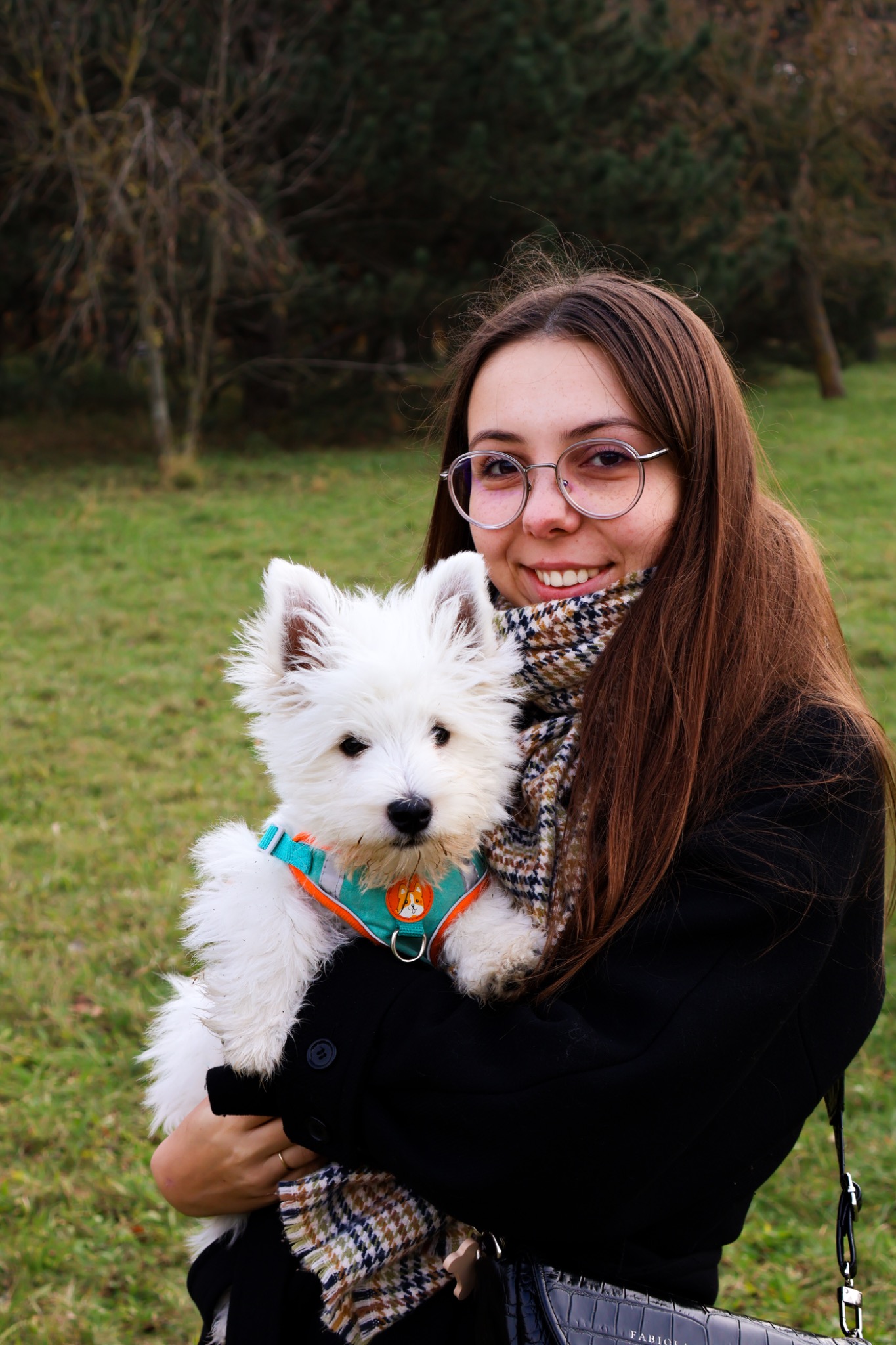 Uśmiechnięta brunetka w okularach trzyma na rękach białego szczeniaka West Highland White Terrier w turkusowo-pomarańczowych szelkach na tle jesiennego parku.