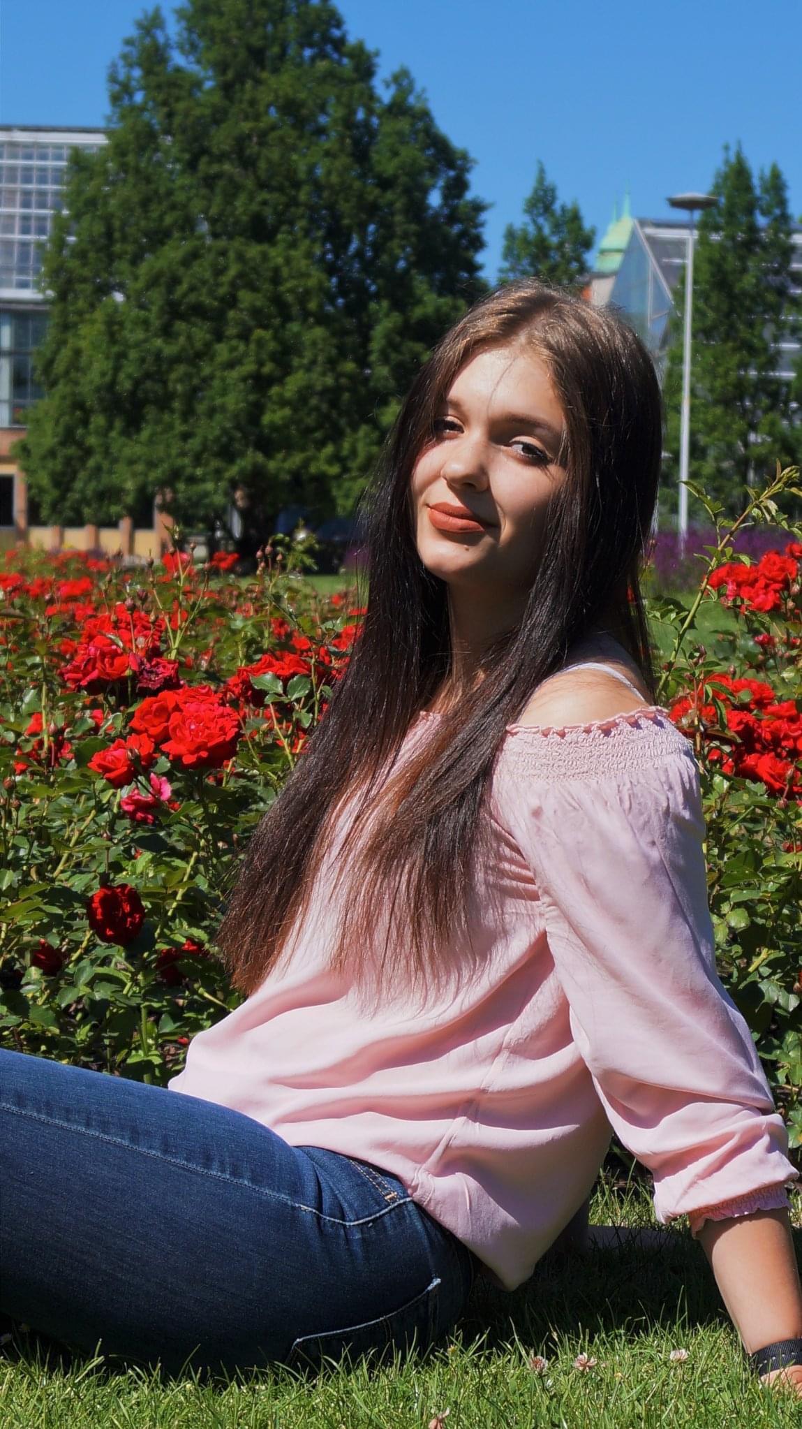 Portret młodej kobiety w różowej bluzce i jeansach, siedzącej na trawie w otoczeniu kwitnących krzewów czerwonych róż, z budynkami i drzewami w tle.