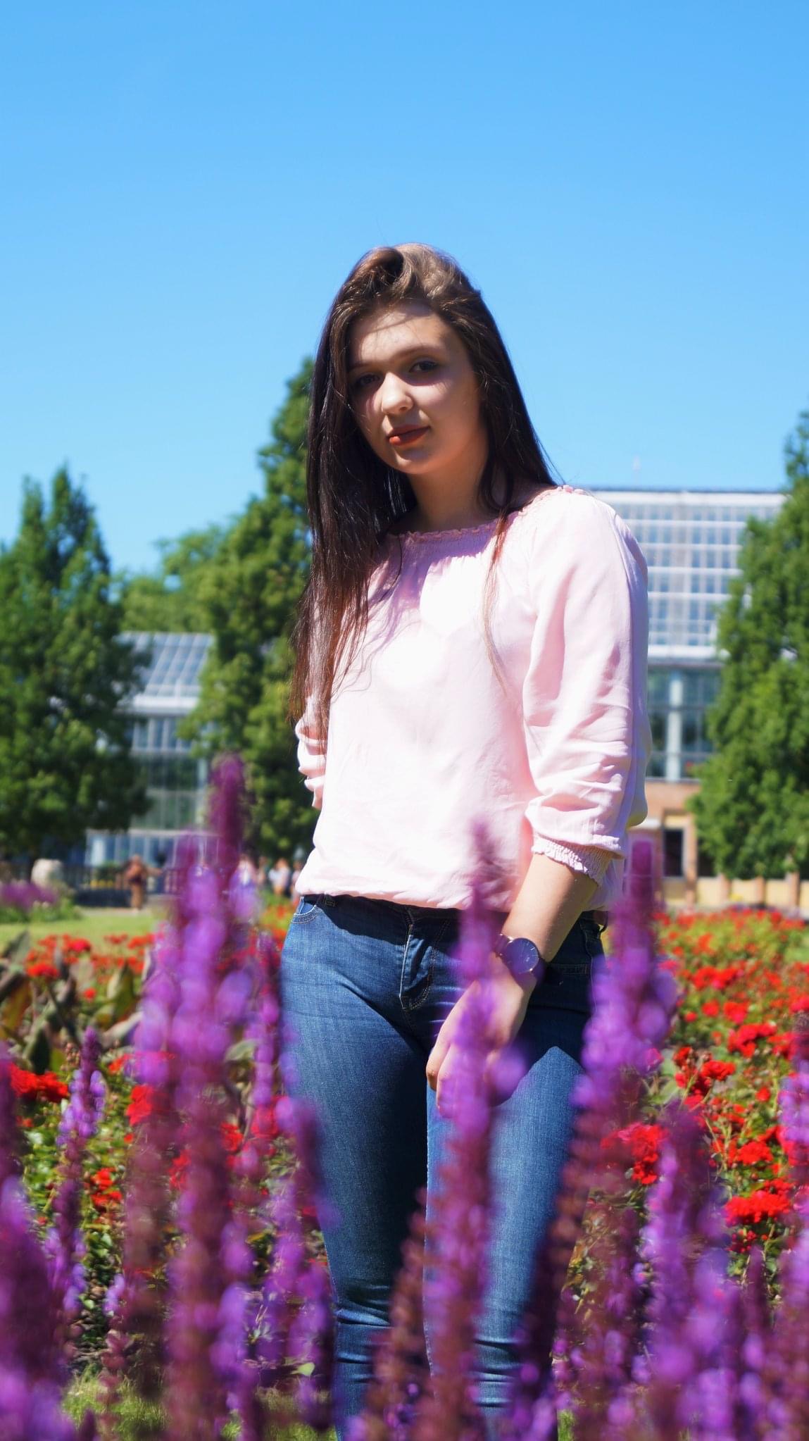 Młoda kobieta w różowej bluzce i jeansach pozuje na tle ogrodu z fioletowymi i czerwonymi kwiatami, w tle szklarnia.