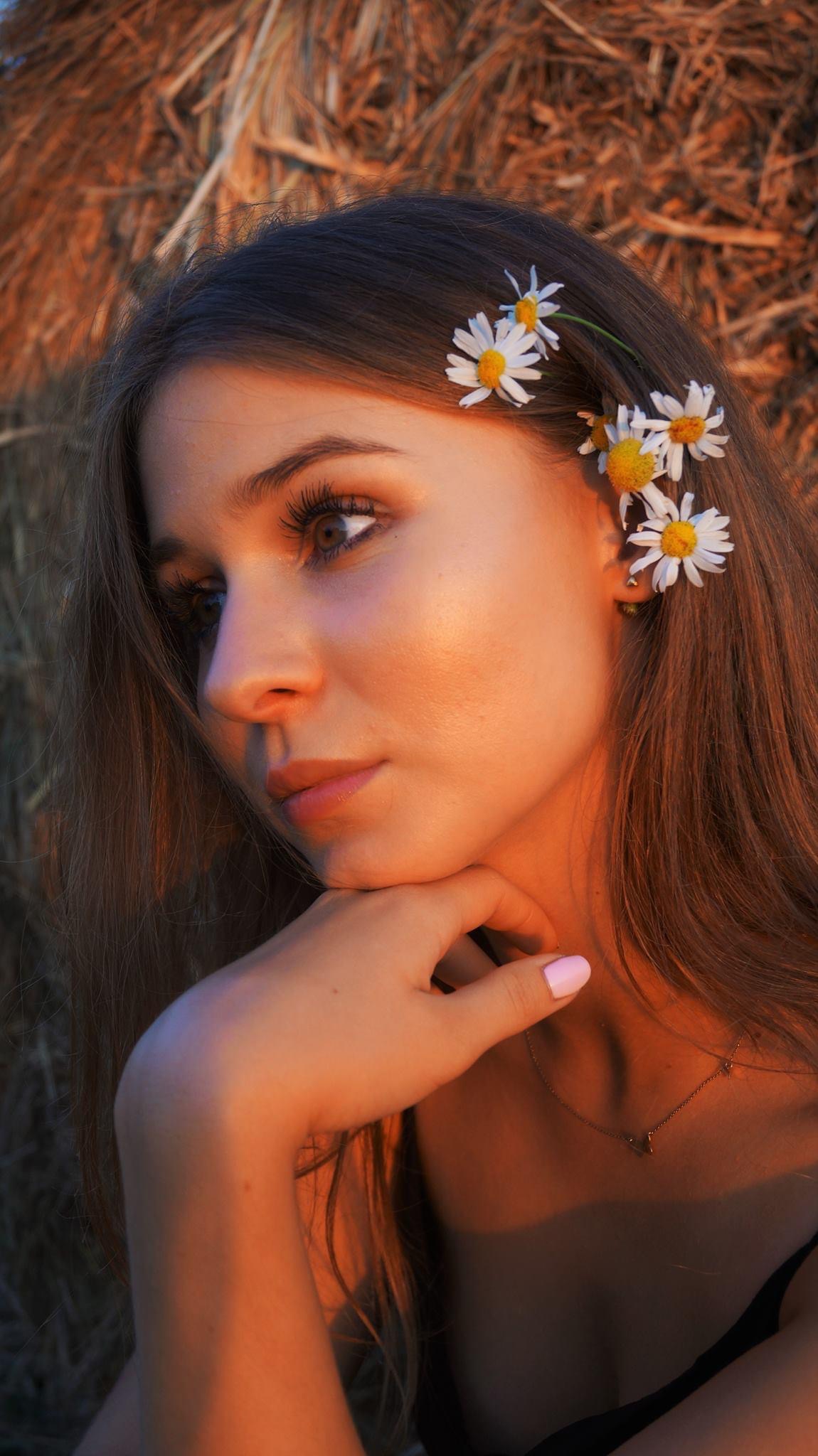 Portret młodej kobiety z brązowymi włosami ozdobionymi białymi stokrotkami, delikatny makijaż, złota biżuteria, w tle stóg siana w ciepłym, zachodzącym słońcu.