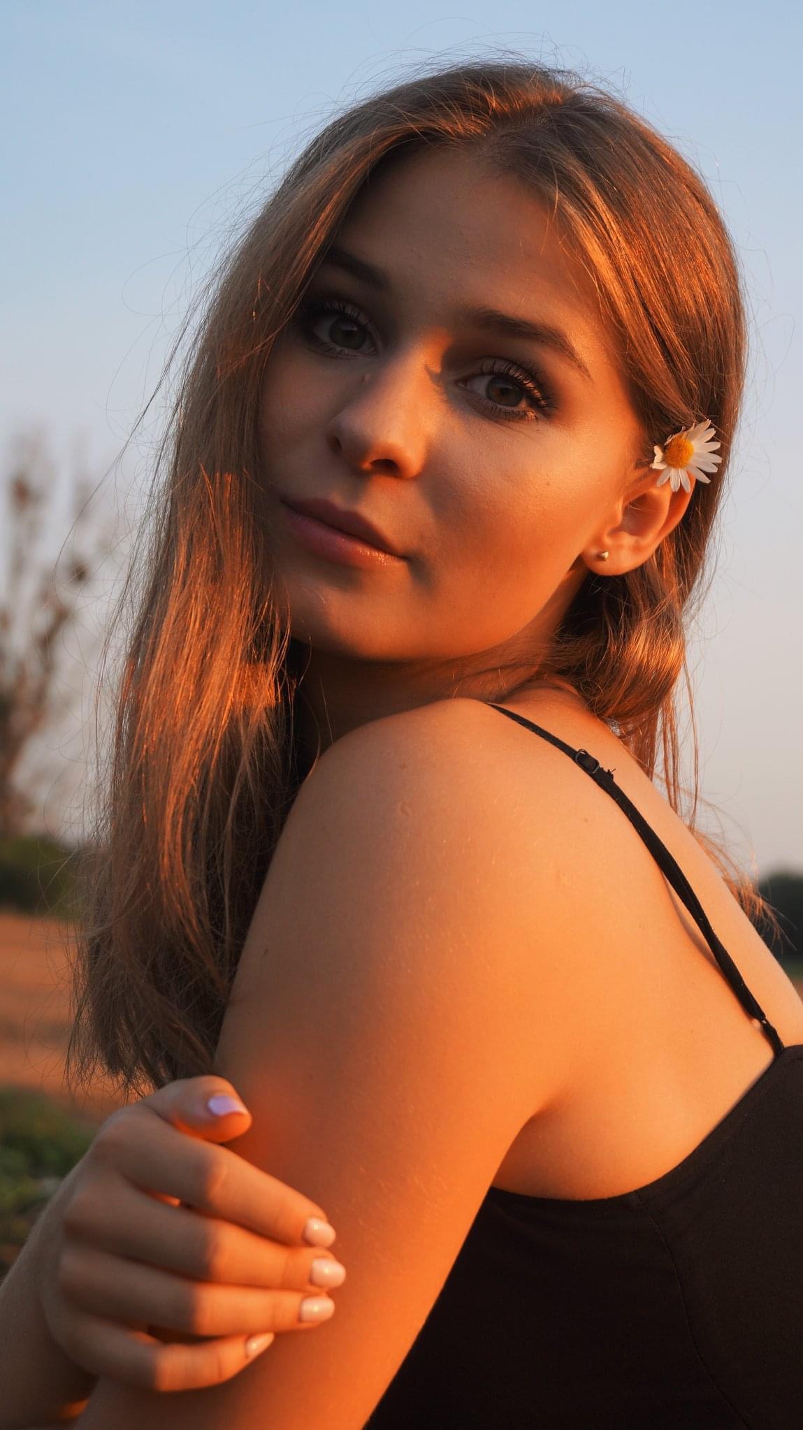 Portret młodej kobiety z rumiankiem we włosach, delikatne światło zachodzącego słońca, czarna sukienka na ramiączkach, naturalny makijaż, lekki uśmiech, pastelowe paznokcie.