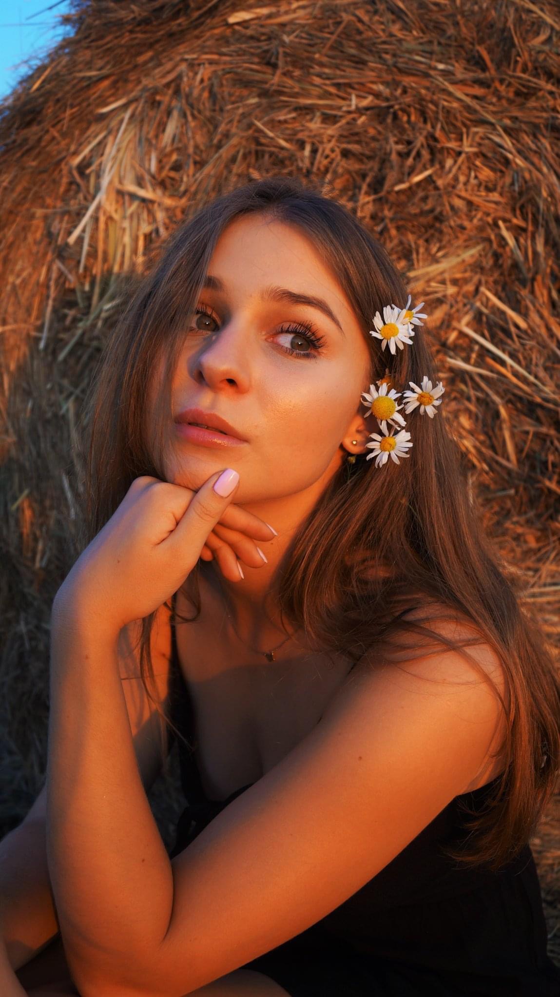 Portret młodej kobiety z rumiankami we włosach, opierającej brodę na dłoni, na tle stogu siana w Rokietnicy, oświetlonej ciepłym światłem zachodzącego słońca.