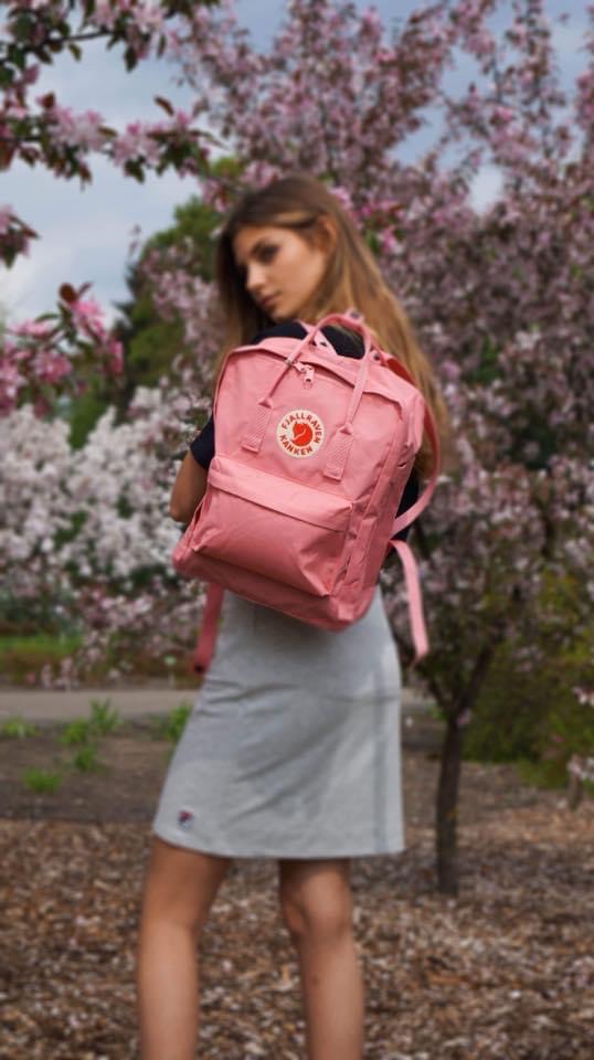 Młoda kobieta z różowym plecakiem Fjallraven Kanken na tle kwitnących drzew wiśni.