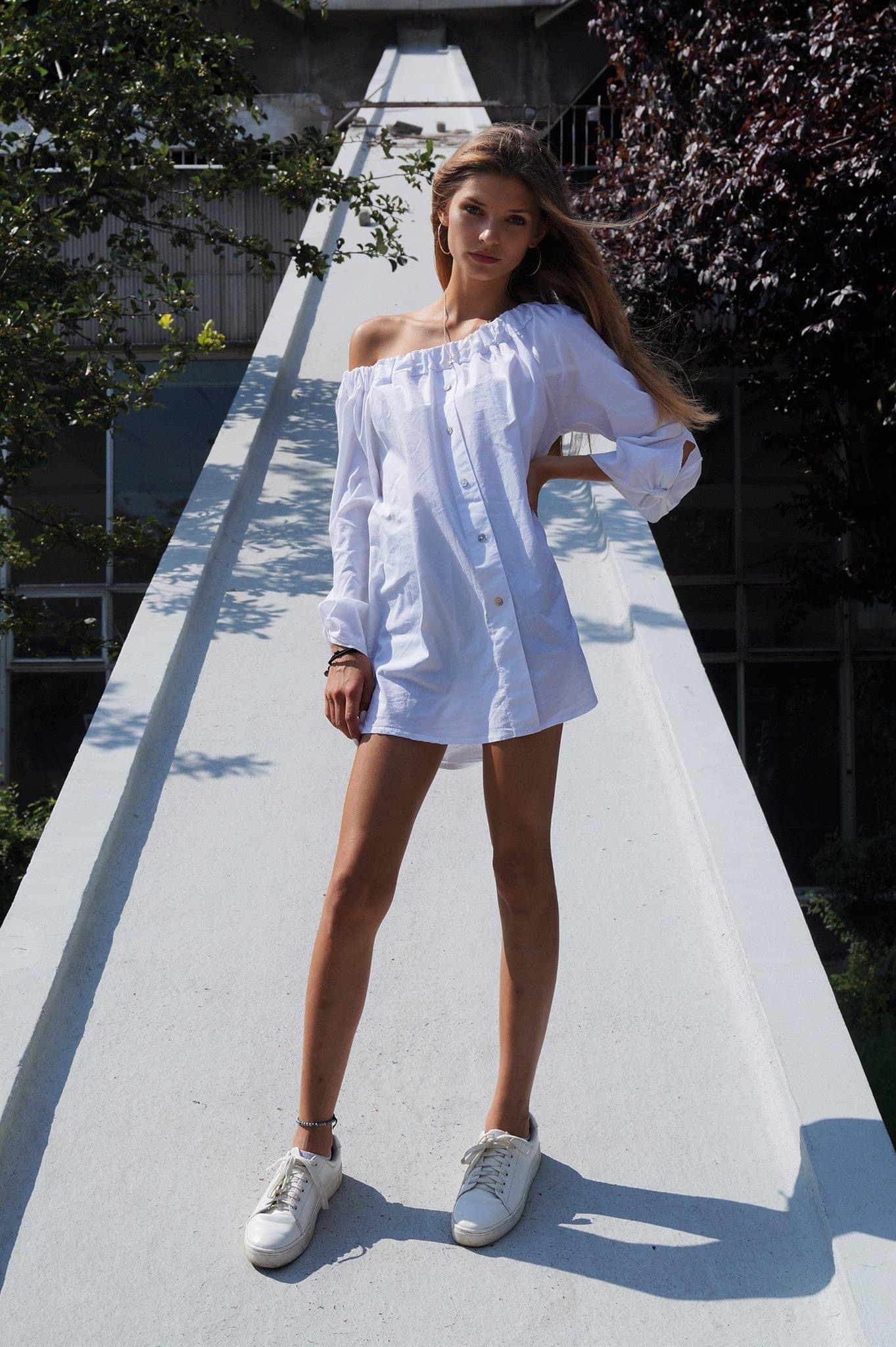 Młoda kobieta w białej, rozpiętej koszuli z odkrytymi ramionami i białych sneakersach pozuje na tle architektonicznym z zielenią.