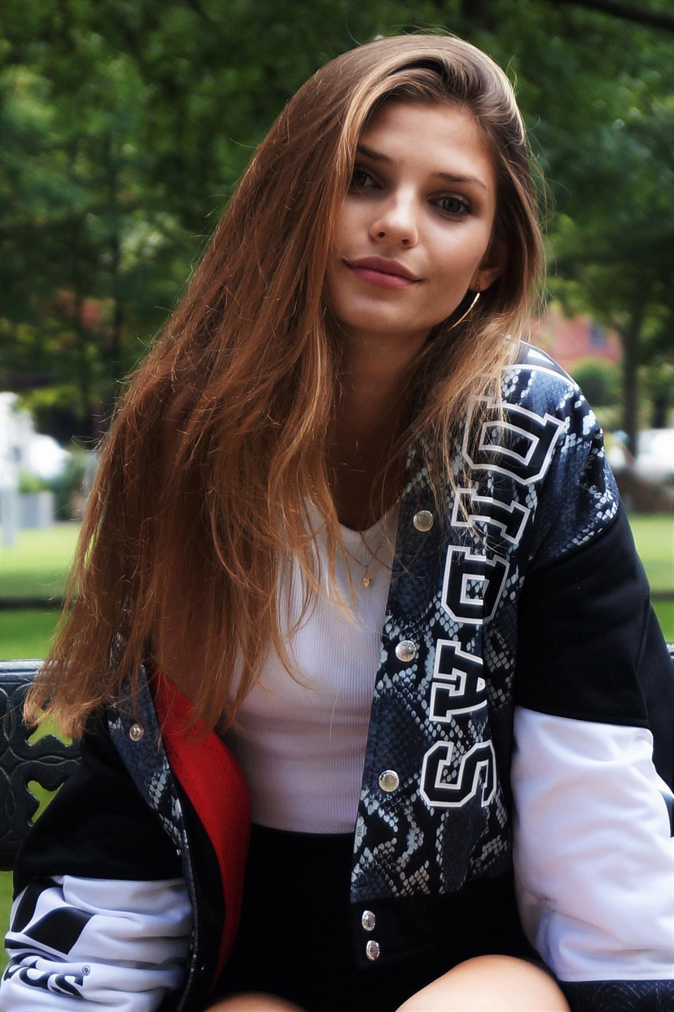 Portret młodej kobiety z długimi brązowymi włosami, ubranej w biały top i kurtkę bomberkę z nadrukiem skóry węża i logo Adidas, siedzącej na ławce w parku.