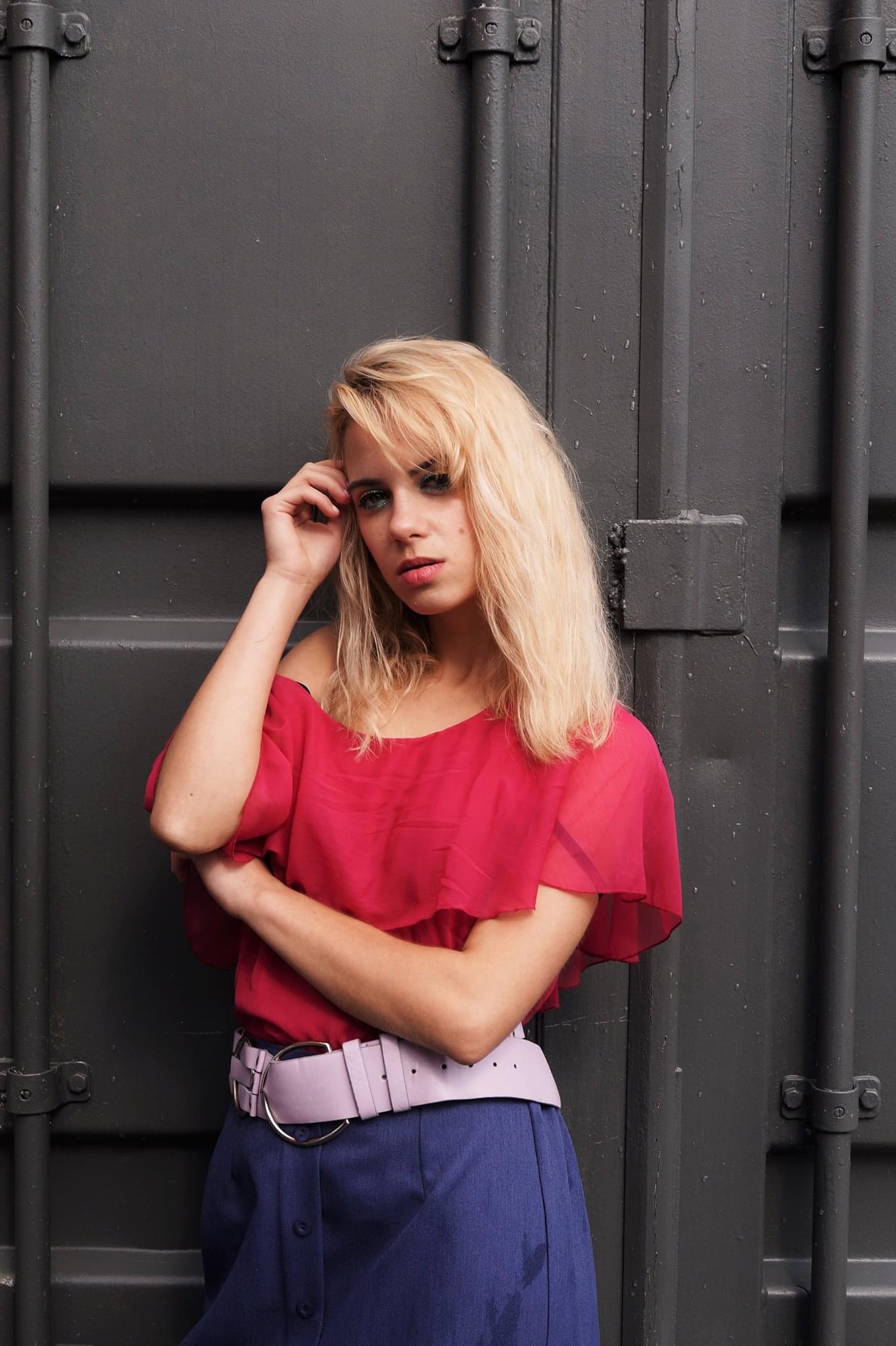 Portret młodej kobiety z blond włosami, w czerwonej bluzce z odkrytymi ramionami i niebieskiej spódnicy, opierającej się o szarą, metalową ścianę.