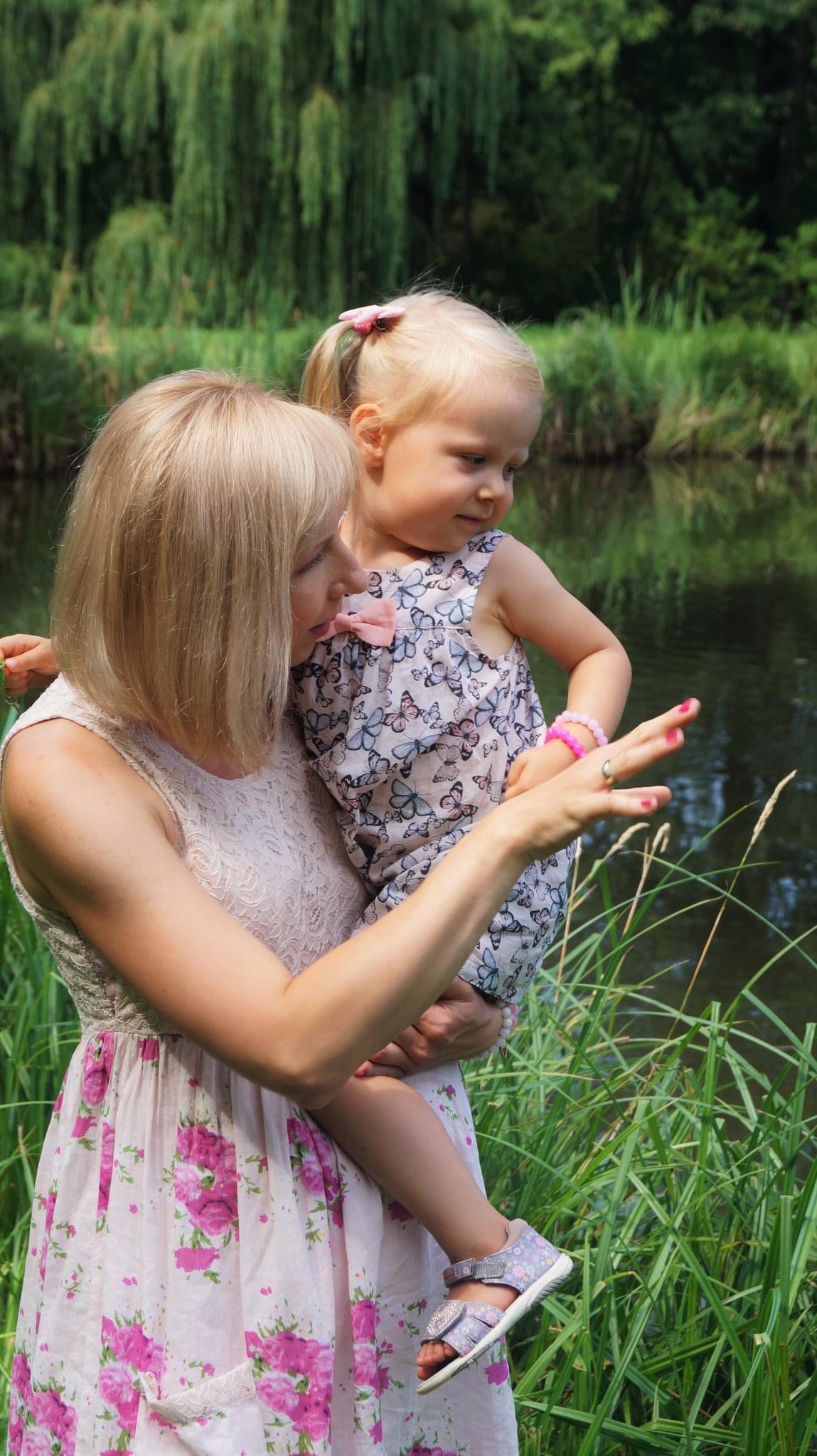 Matka trzyma córkę nad brzegiem jeziora, obie ubrane w letnie sukienki w kwiaty i motyle, dziewczynka z różową spinką we włosach i bransoletką na ręce, wskazują palcami na coś w trawie.