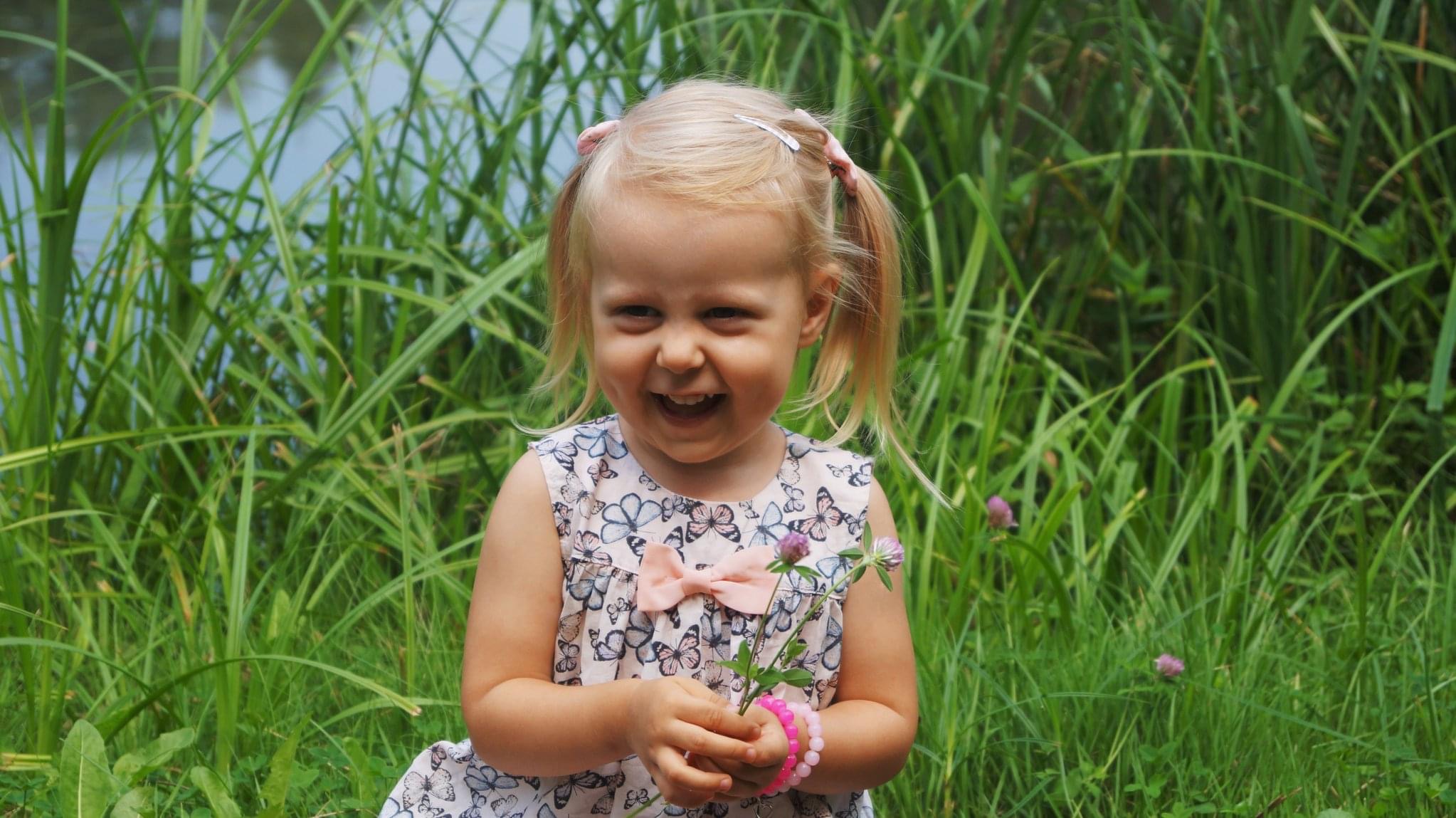 Uśmiechnięta blond dziewczynka w sukience z motylami, trzymająca koniczynę i różową bransoletkę, siedząca na tle zieleni i wody.