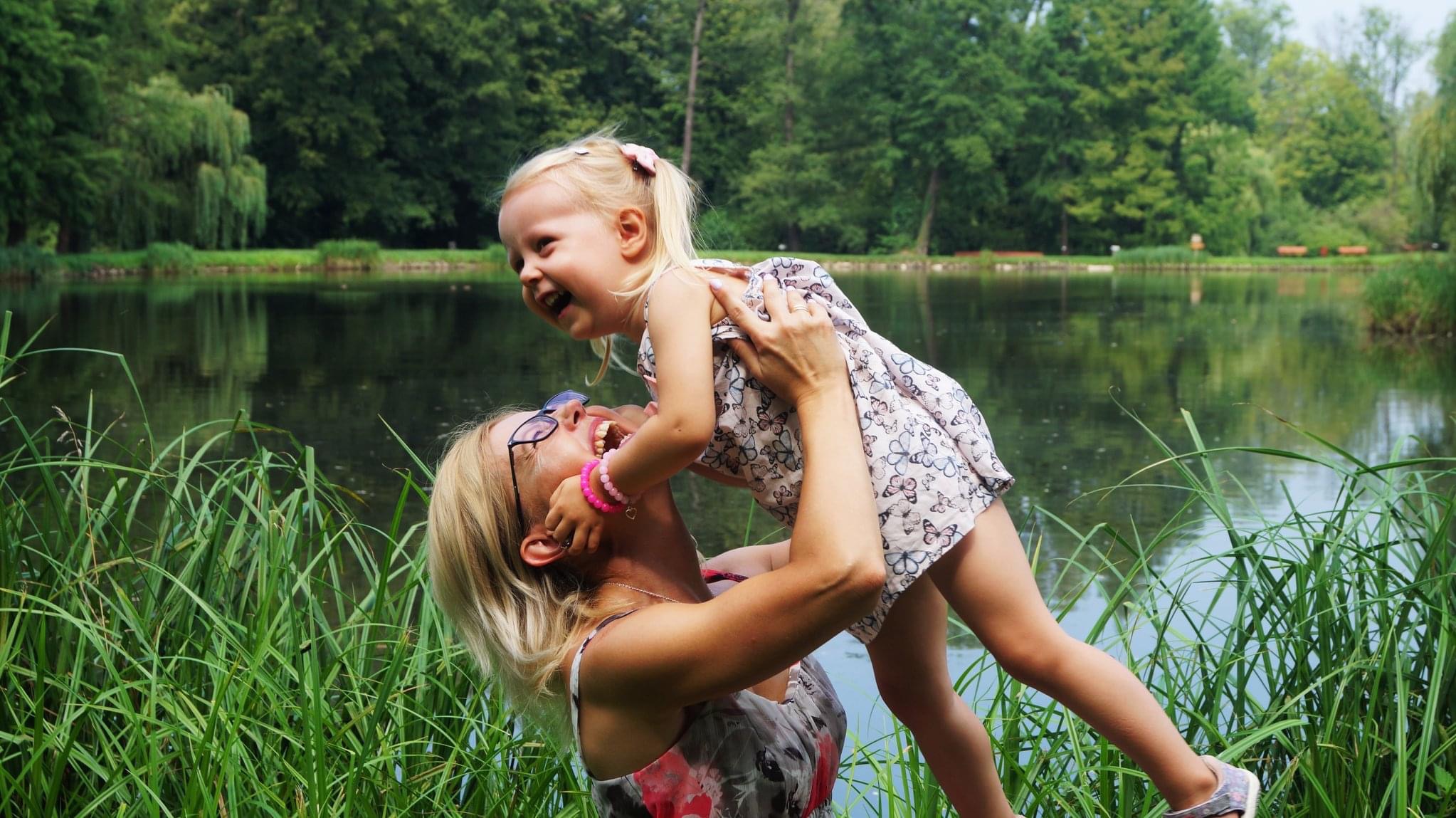 Uśmiechnięta kobieta w okularach podnosi w górę roześmianą blondwłosą dziewczynkę w sukience z motylami na tle jeziora i zieleni.