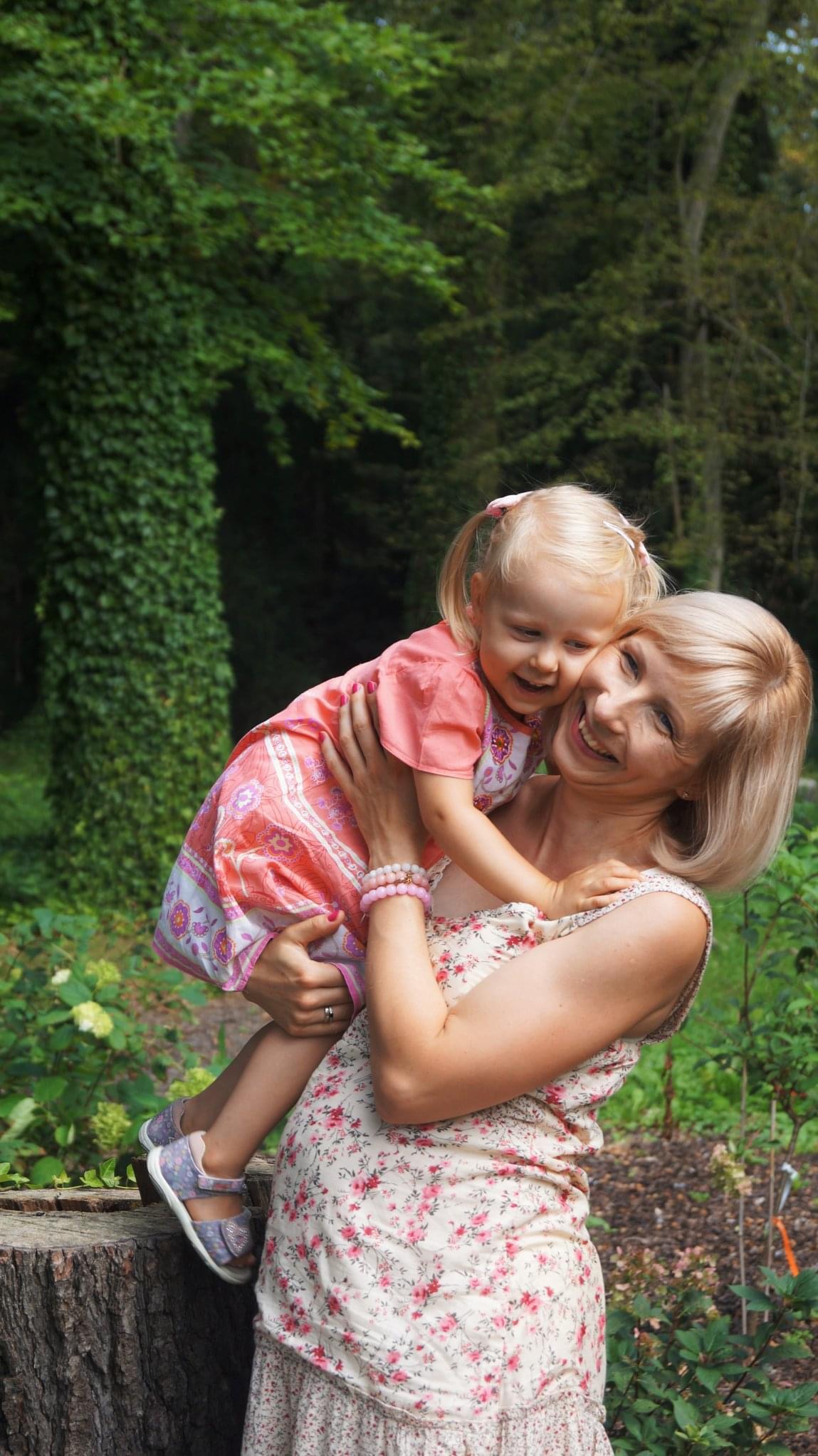 Uśmiechnięta blond mama trzymająca na rękach roześmianą córeczkę w różowej sukience, na tle zieleni drzew i pnia.