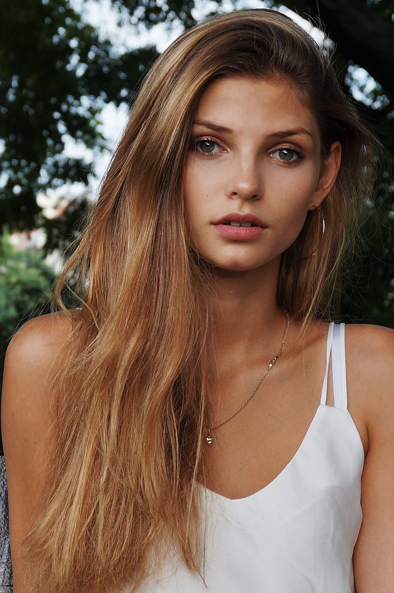 Portret młodej kobiety z długimi blond włosami, noszącej biały top na ramiączkach i złotą biżuterię, na tle zieleni.