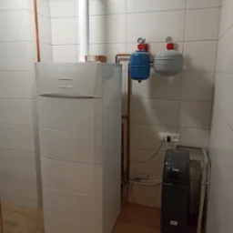 Nowoczesny, biały piec kondensacyjny z podłączonymi instalacjami miedzianymi i zbiornikami wyrównawczymi w pomieszczeniu wyłożonym białymi płytkami, widoczny także filtr do wody.