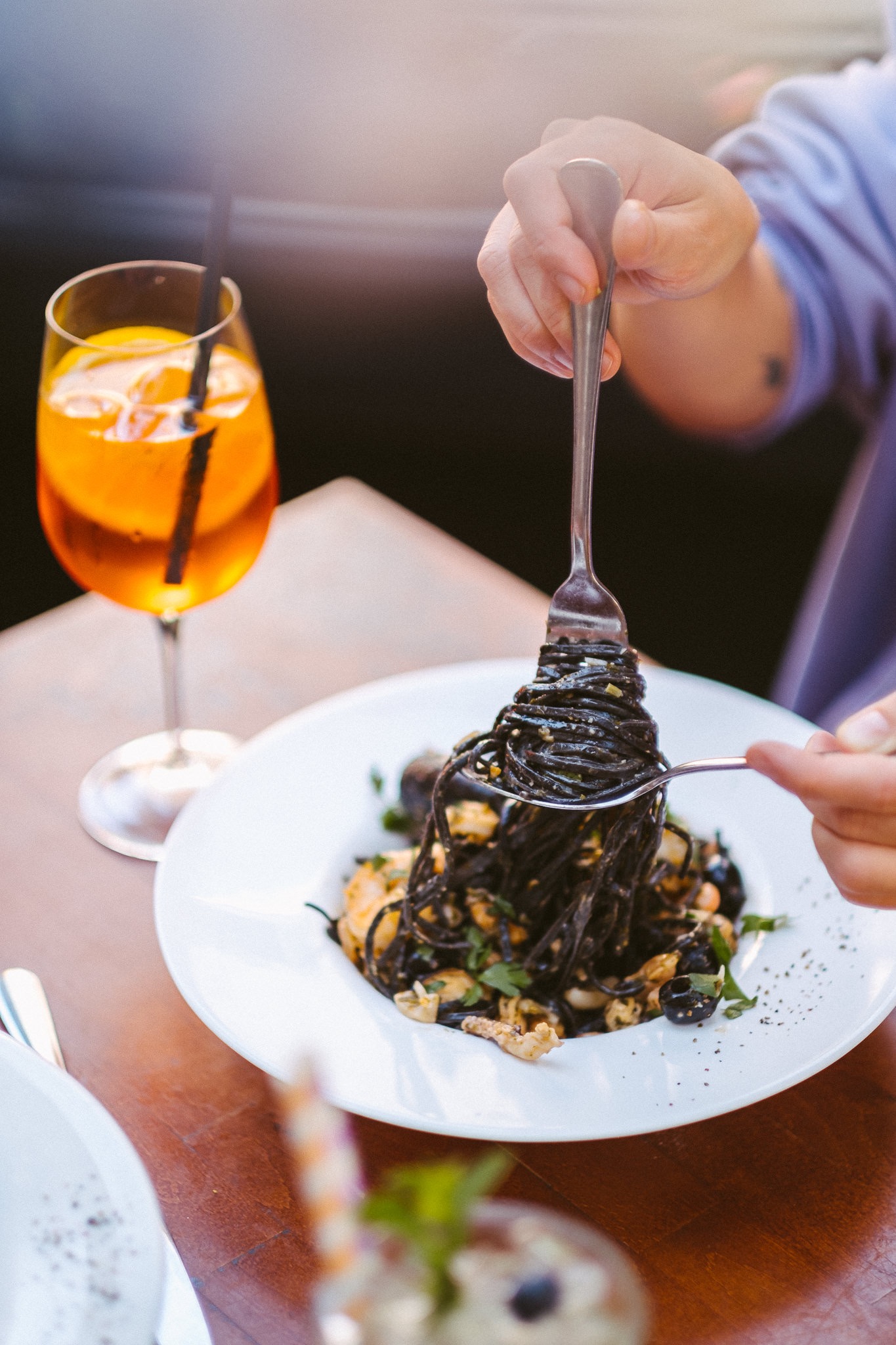 Czarny makaron z owocami morza podawany na białym talerzu w Trattoria Corleone, podnoszony widelcem, obok drink Aperol Spritz.