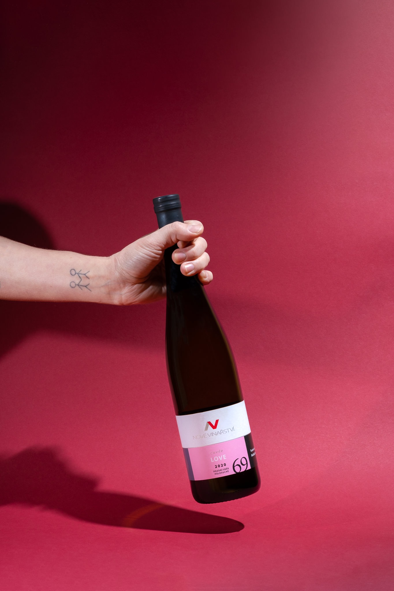Dłoń z tatuażem trzyma butelkę wina Nové Vinarstvi Cuvée Love 2020 na burgundowym tle, sesja produktowa dla Winny Winiarnia.