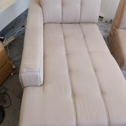 Kacper Bakuła Diamond Professional Cleaning - Jasna, beżowa kanapa z poduchami i przeszyciami, widoczna z góry, z fragmentem otoczenia: szara wykładzina, biała ściana i fragment mebla z jasnego drewna.