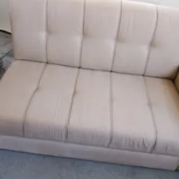 Jasnobeżowa, dwuosobowa kanapa z pikowanym oparciem i charakterystycznymi pionowymi przeszyciami, ustawiona na szarym dywanie.