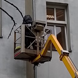 Kacper Bakuła Diamond Professional Cleaning - Pracownik w podnośniku koszowym, ubrany w ciemną odzież ochronną, przy ścianie budynku z oknami. Żółte ramię podnośnika widoczne z prawej strony.