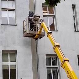 Kacper Bakuła Diamond Professional Cleaning - Pracownik w podnośniku koszowym na tle szarej ściany budynku z oknami, prawdopodobnie wykonujący prace na wysokościach.