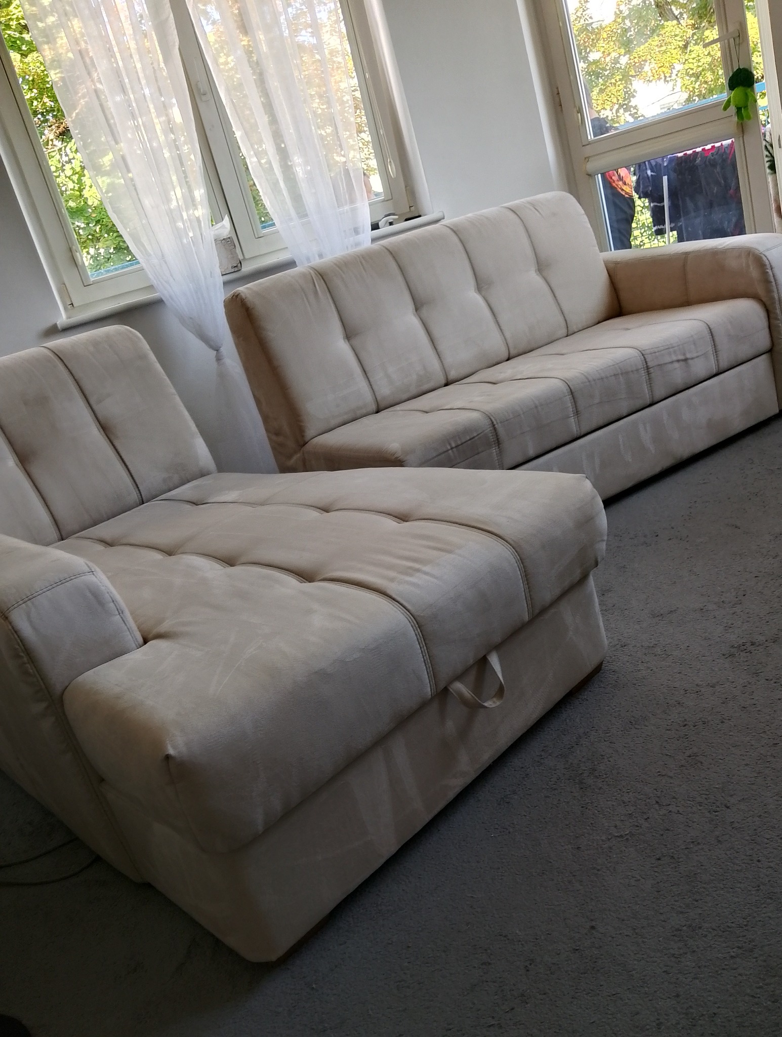 Jasna, tapicerowana sofa i narożnik z widocznymi przeszyciami i guzikami na siedziskach, ustawione w salonie z oknami zasłoniętymi białymi firankami.