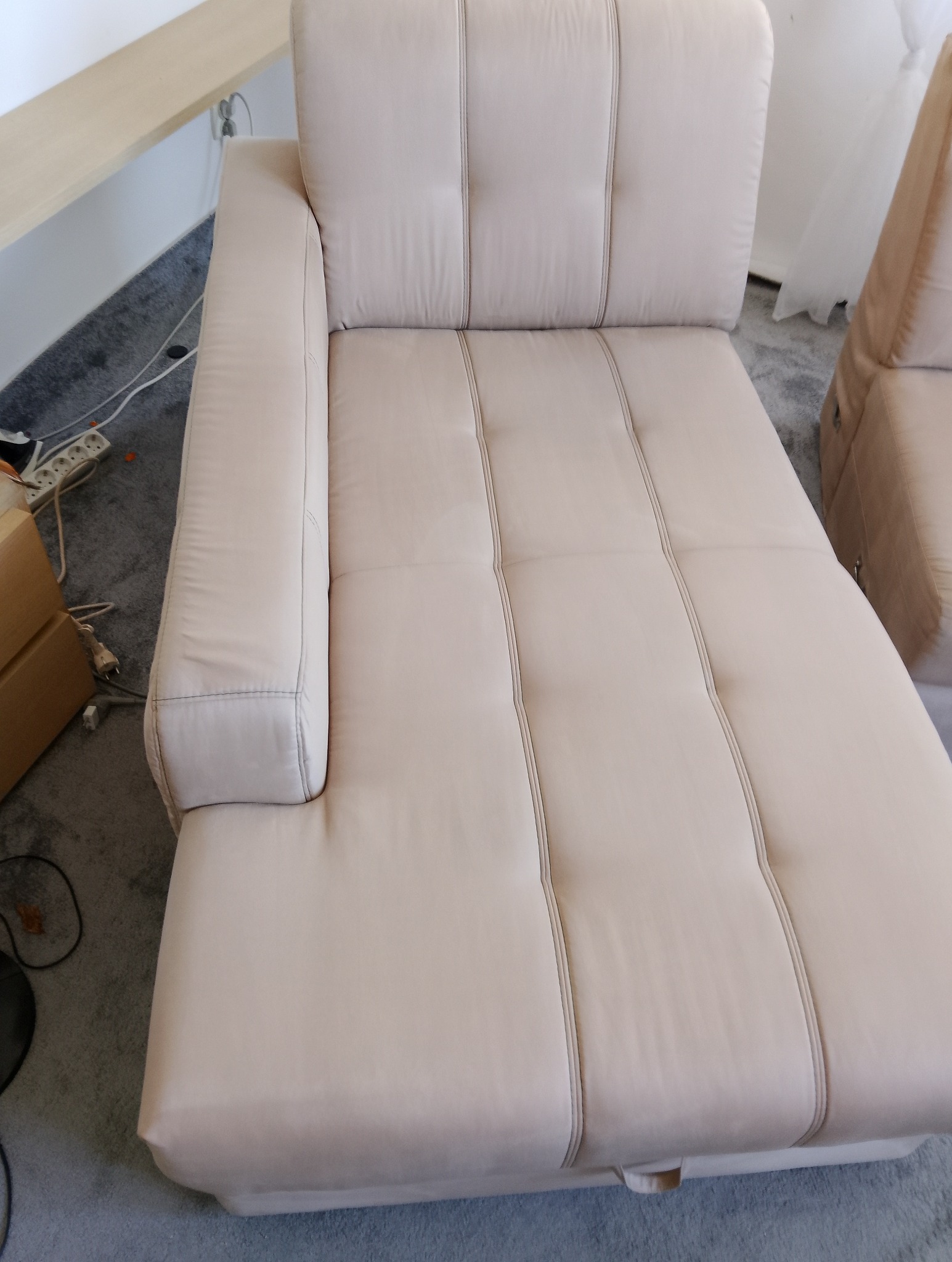 Jasna, beżowa kanapa z poduchami i przeszyciami, widoczna z góry, z fragmentem otoczenia: szara wykładzina, biała ściana i fragment mebla z jasnego drewna.