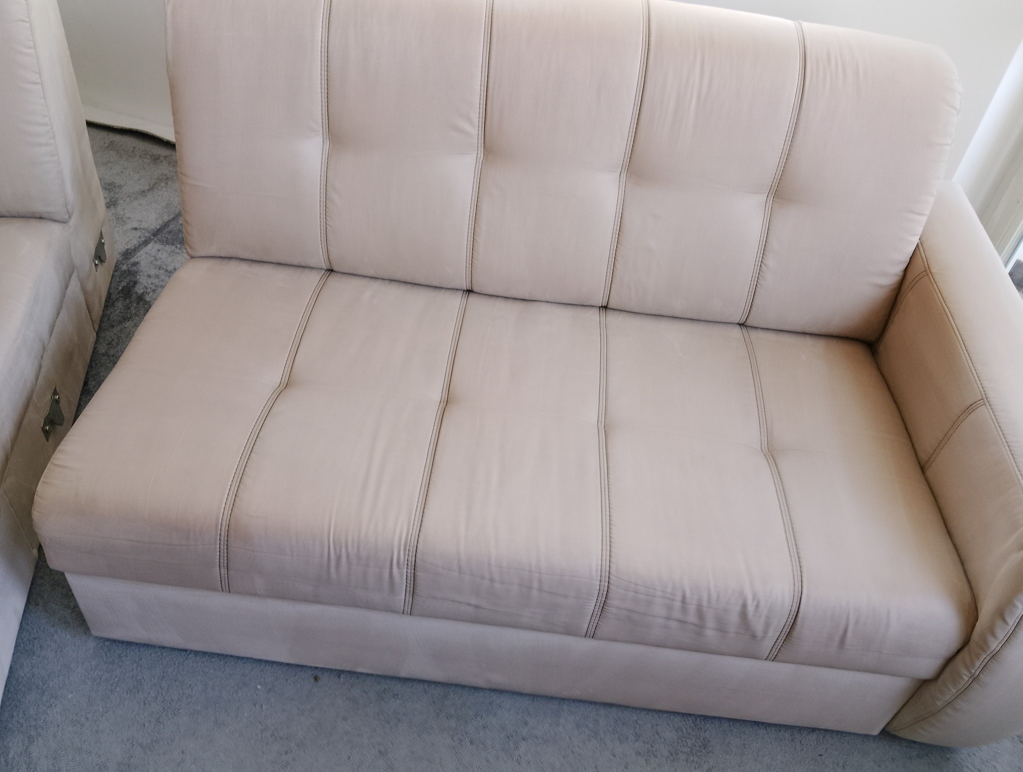 Jasnobeżowa, dwuosobowa kanapa z pikowanym oparciem i charakterystycznymi pionowymi przeszyciami, ustawiona na szarym dywanie.