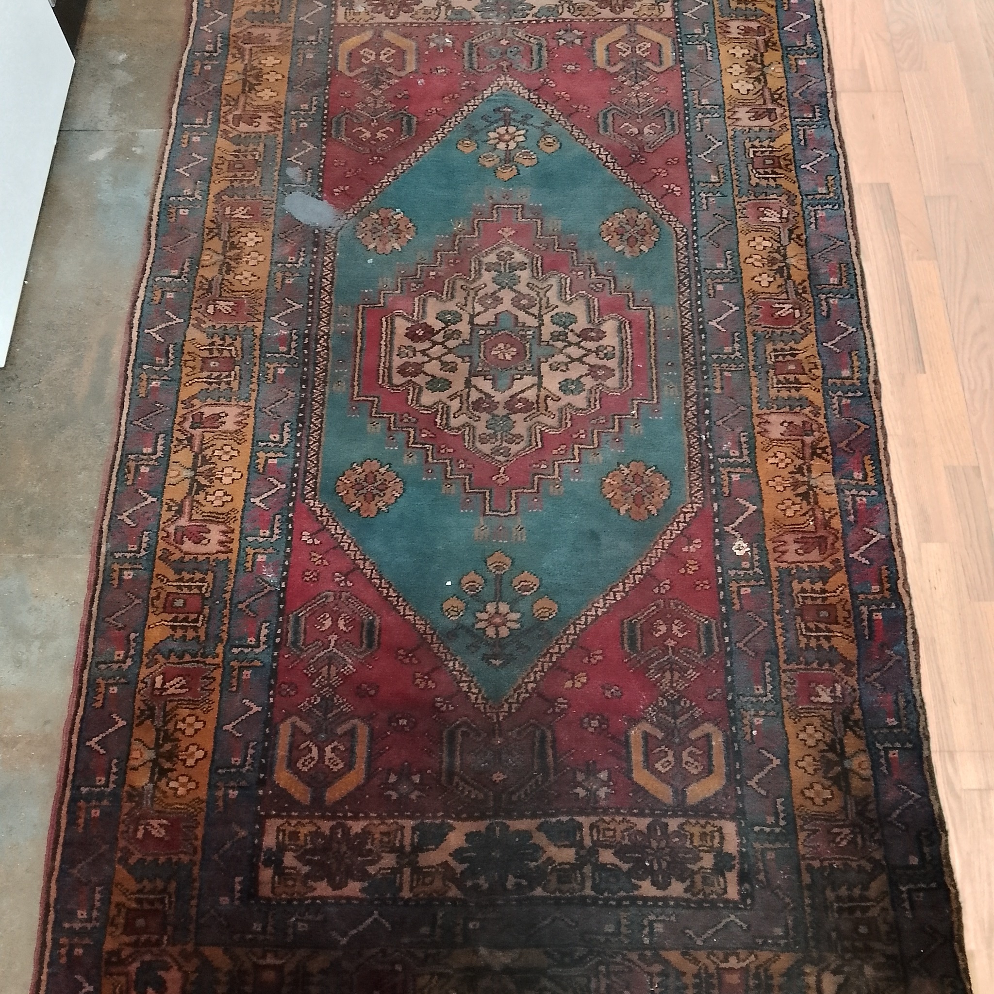 Wzorzysty, orientalny dywan w odcieniach czerwieni, turkusu i brązu leżący na podłodze z jasnych paneli, widoczne lekkie ślady użytkowania i przebarwienia.