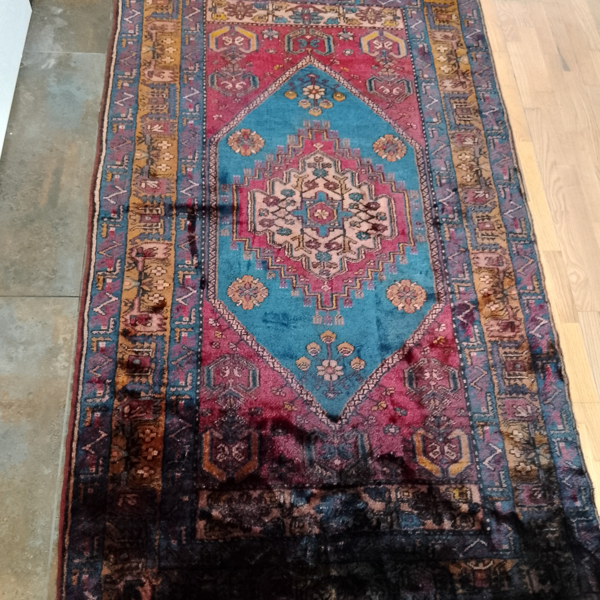 Wzorzysty, orientalny dywan w odcieniach czerwieni, błękitu i brązu, leżący na podłodze, z widocznymi śladami użytkowania i potencjalnymi zabrudzeniami w dolnej części.