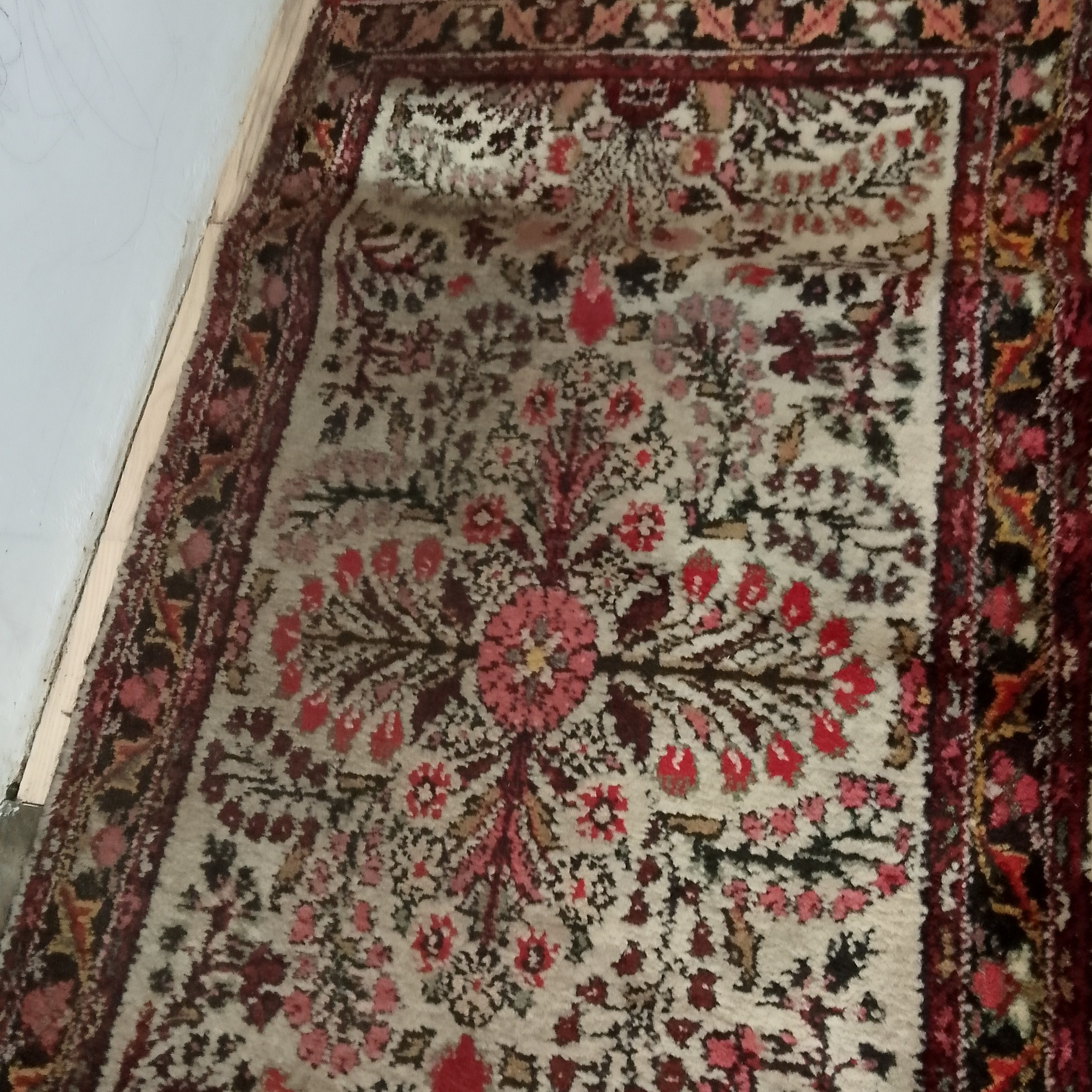 Wzorzysty, orientalny dywan z czerwono-różowymi motywami kwiatowymi, leżący na podłodze przy białej ścianie.