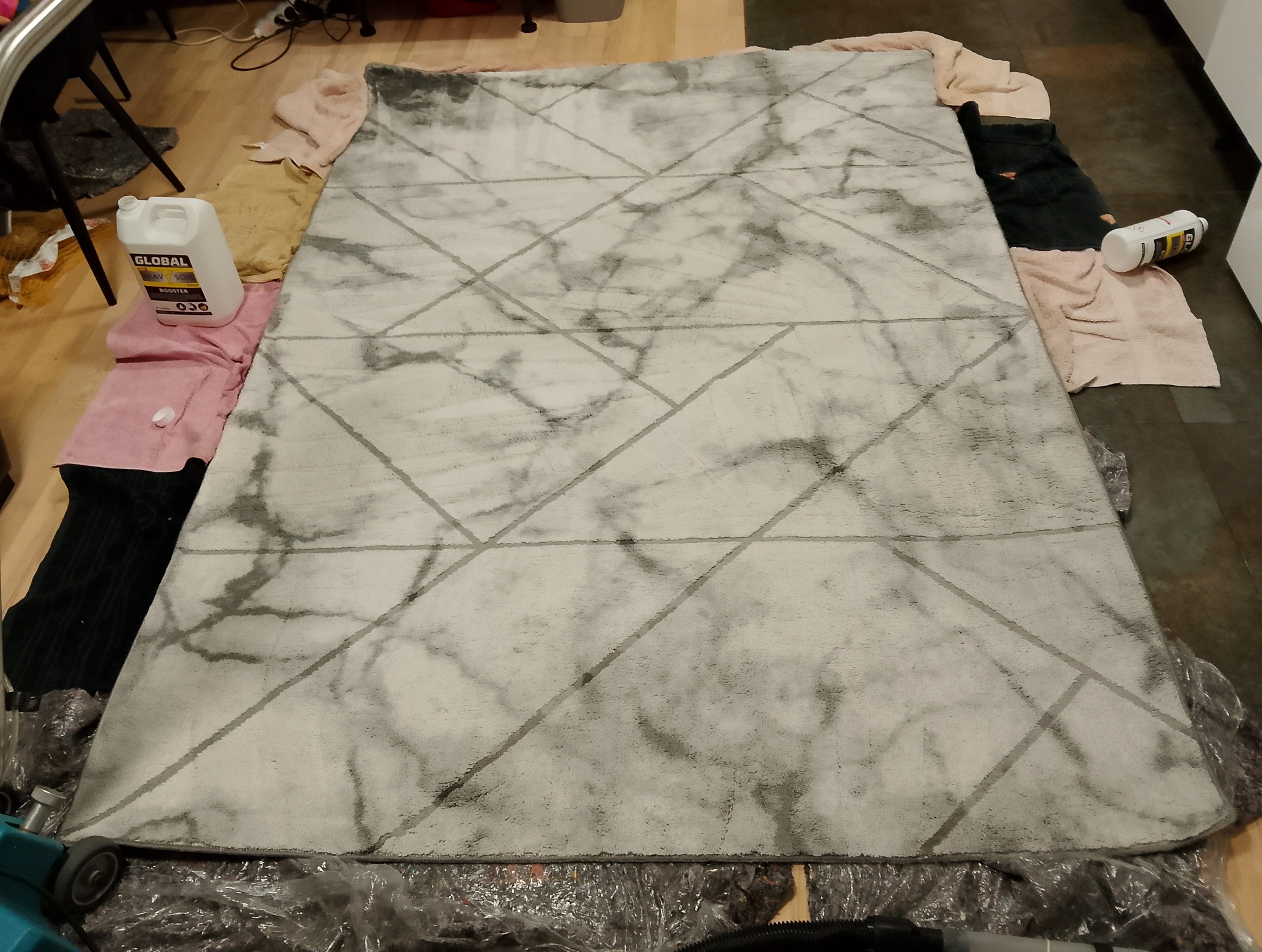 Biało-szary dywan z geometrycznym wzorem, otoczony ręcznikami i środkami czyszczącymi, przygotowany do prania.