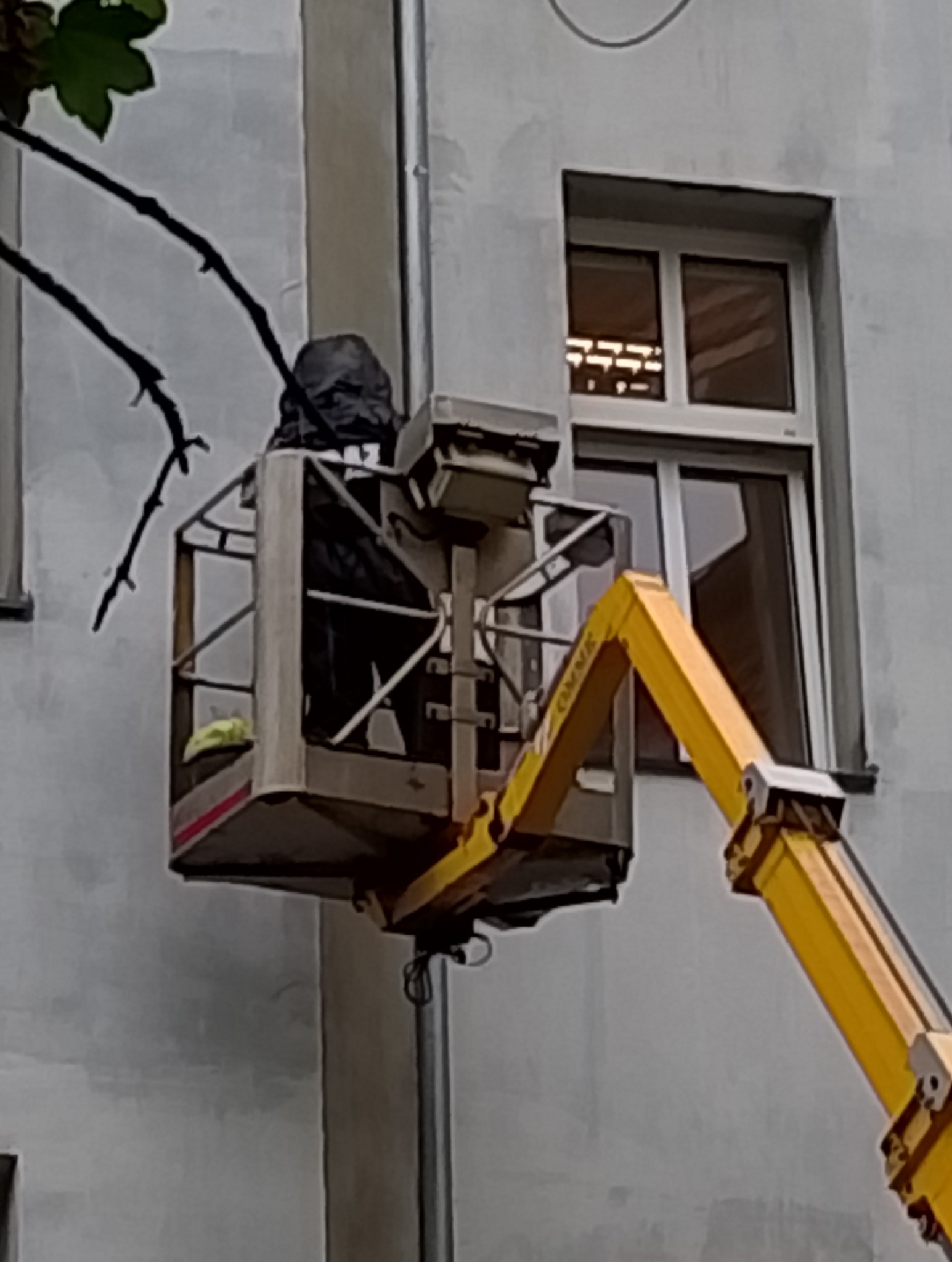 Pracownik w podnośniku koszowym, ubrany w ciemną odzież ochronną, przy ścianie budynku z oknami. Żółte ramię podnośnika widoczne z prawej strony.
