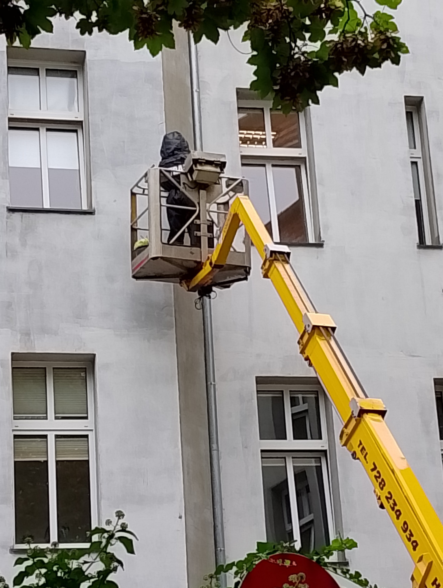 Pracownik w podnośniku koszowym na tle szarej ściany budynku z oknami, prawdopodobnie wykonujący prace na wysokościach.