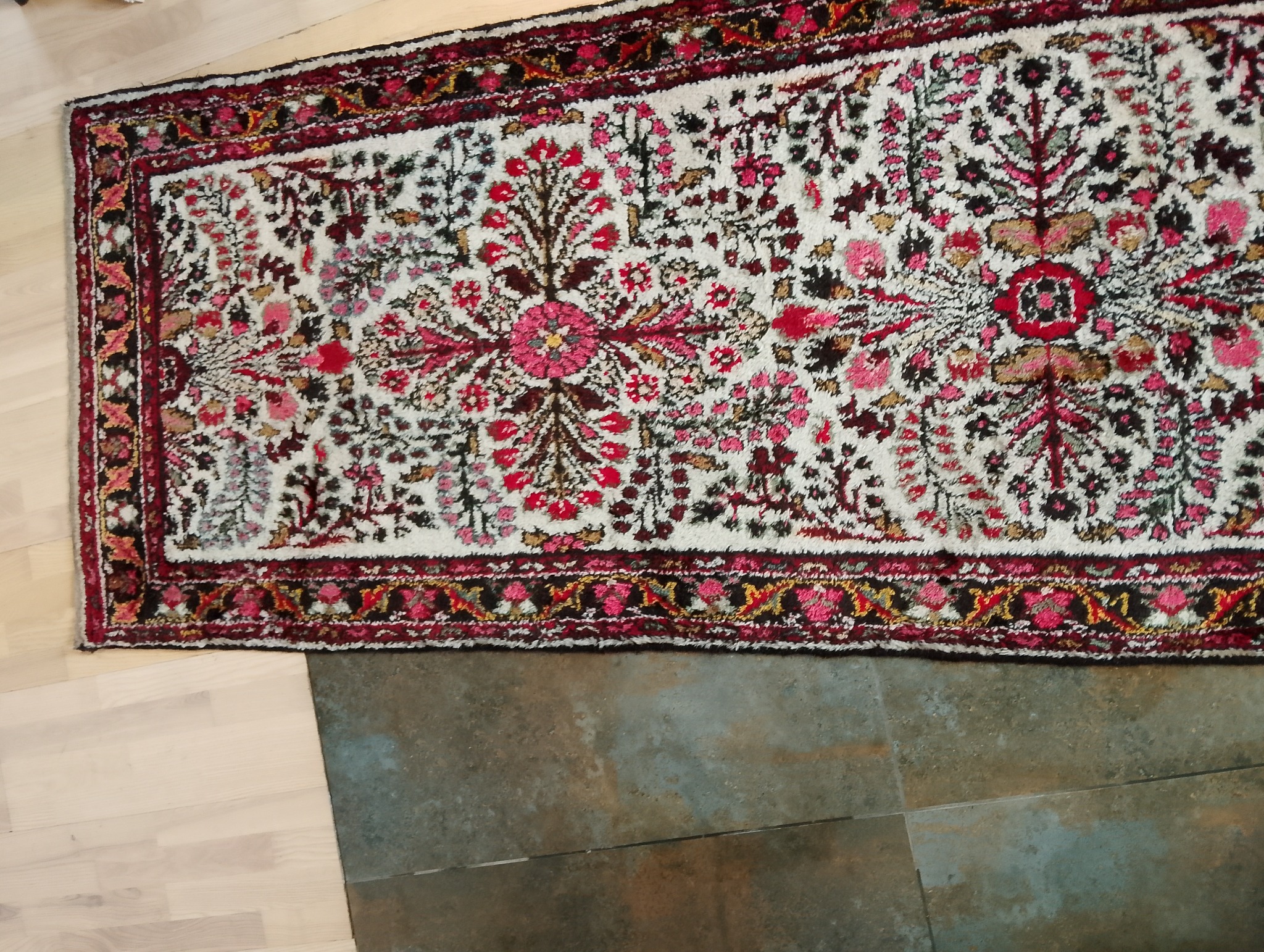 Fragment wzorzystego dywanu orientalnego z motywami kwiatowymi w odcieniach czerwieni, różu i czerni na białym tle, leżącego na granicy drewnianej podłogi i szarej, matowej płytki.