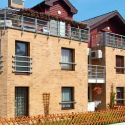 Kompleks budynków mieszkalnych z cegły klinkierowej, balkony z metalowymi balustradami, kwiaty w donicach na balkonach i przy ogrodzeniu, zadbany trawnik i oświetlenie uliczne.