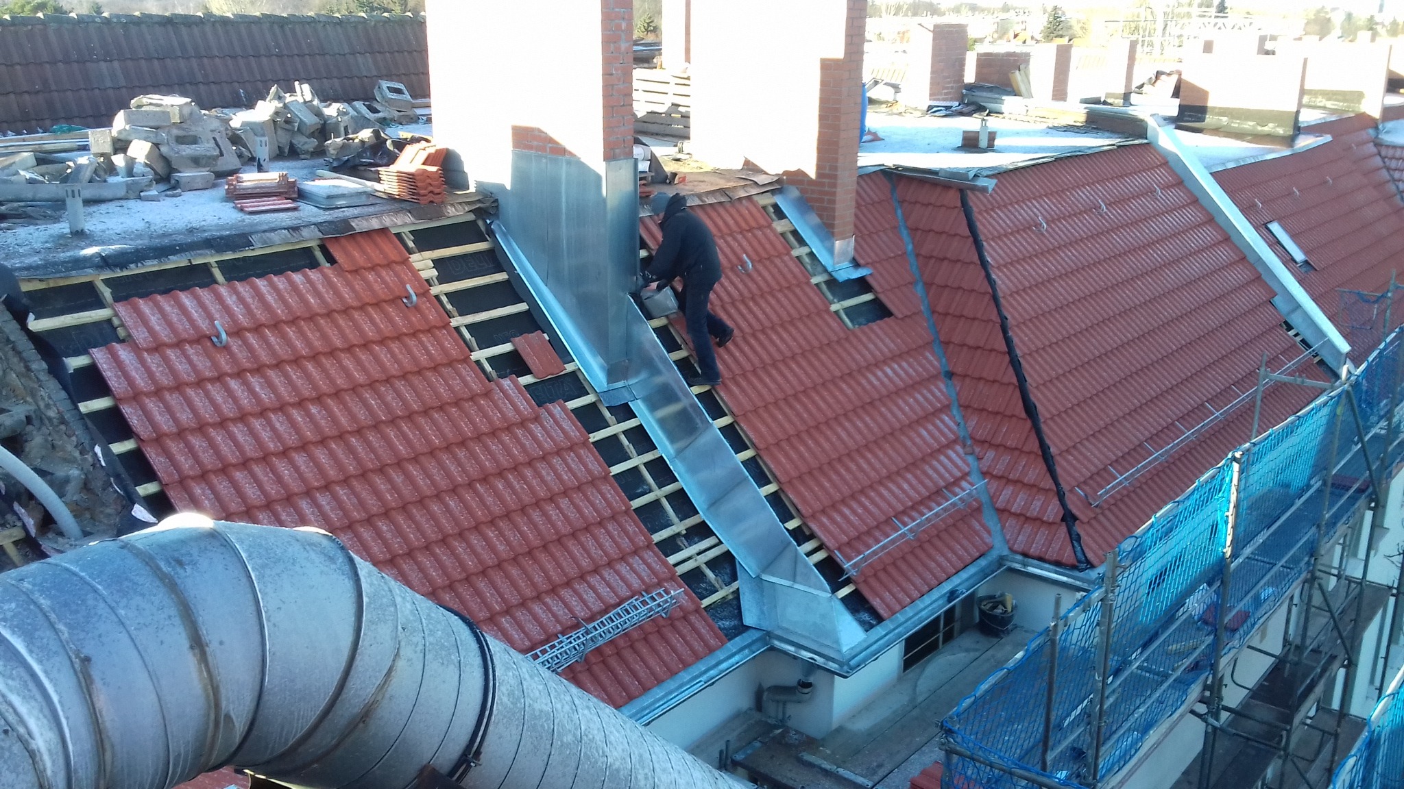 Prace dekarskie na dachu z czerwonej dachówki, montaż obróbek blacharskich komina przez dekarza, widoczne elementy konstrukcji dachu i rusztowania.