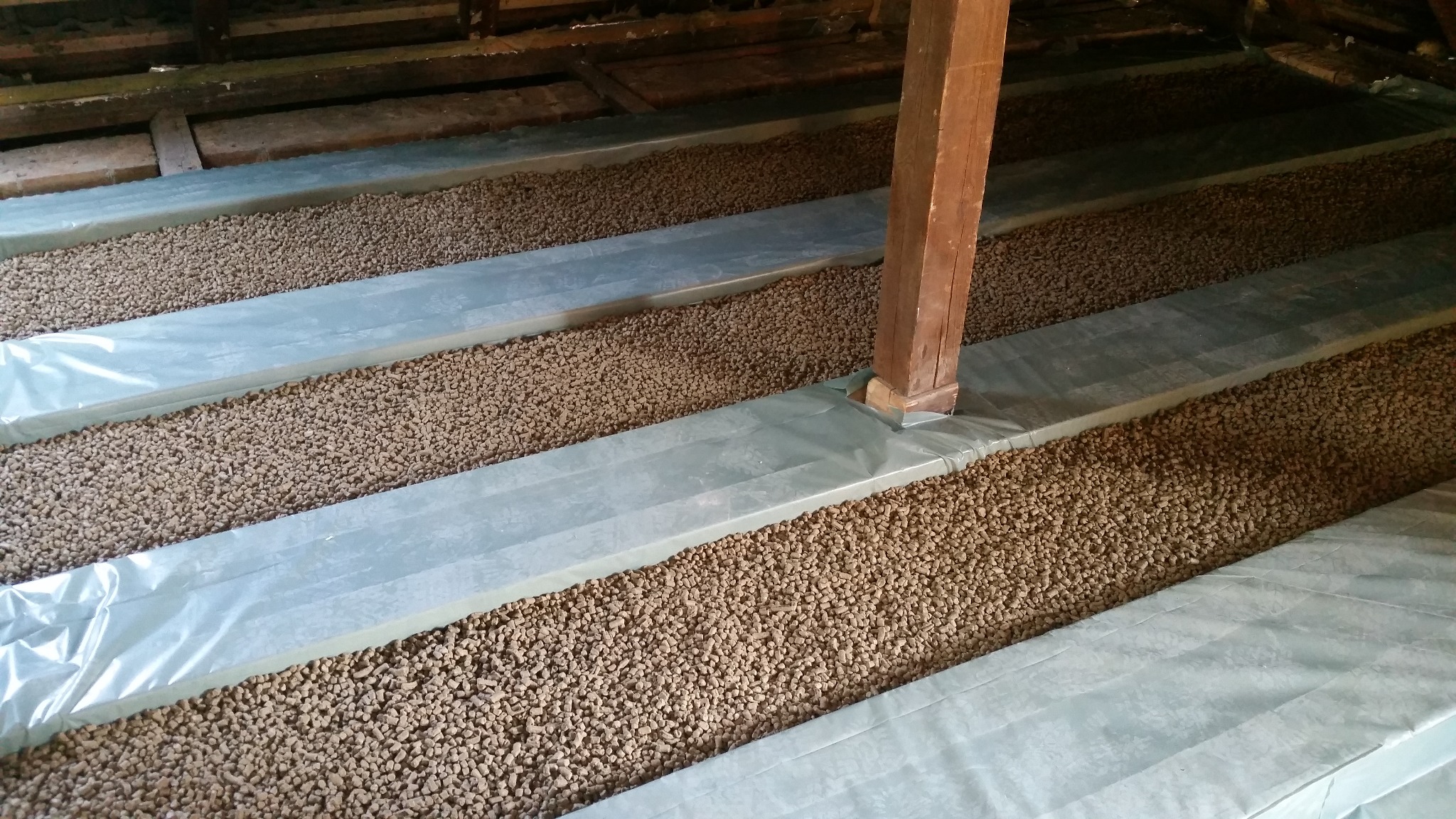 Izolacja poddasza z użyciem granulatu, zabezpieczona folią, widoczny drewniany słup konstrukcyjny.