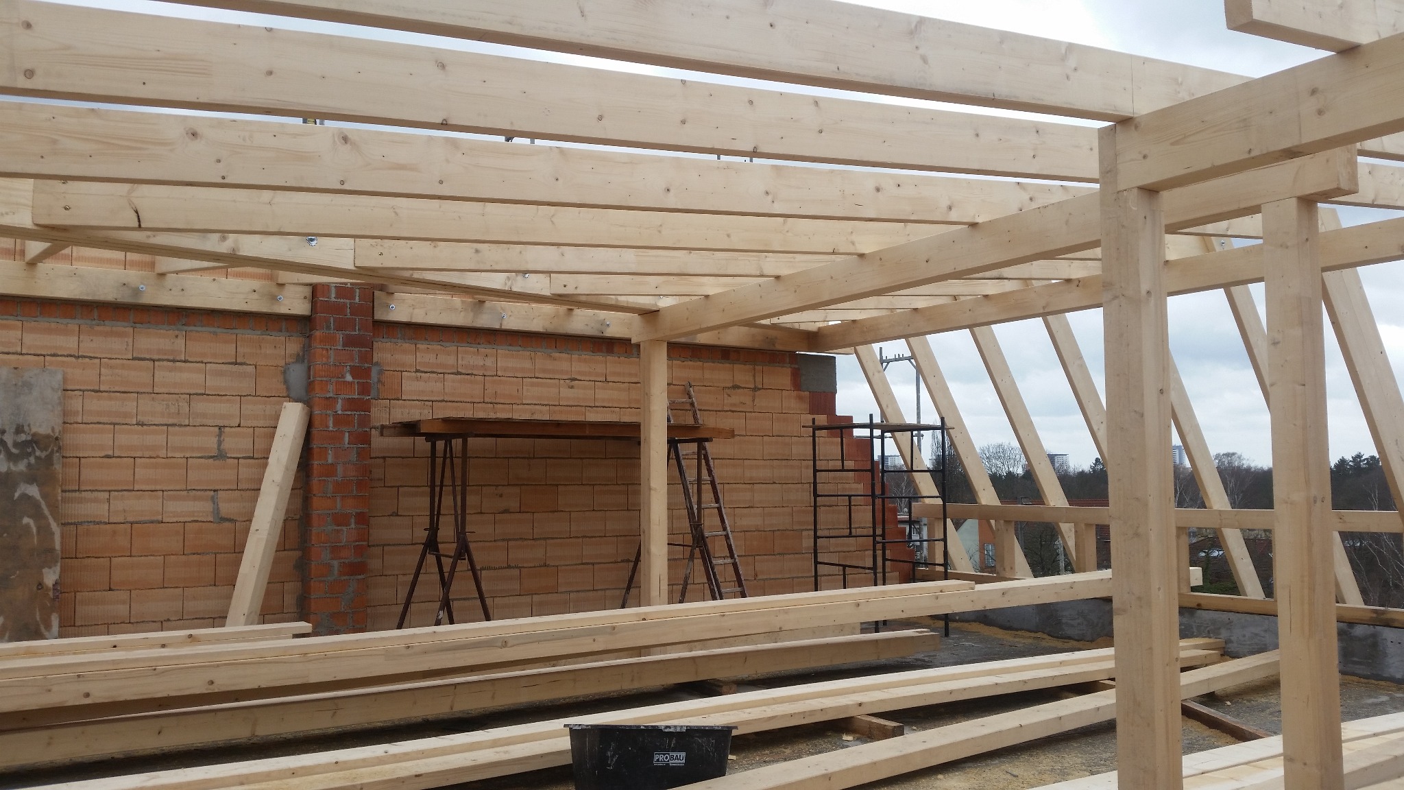 Konstrukcja drewniana dachu w trakcie budowy, widoczne belki, słupy, mur z cegły dziurawki, rusztowania i drabiny.