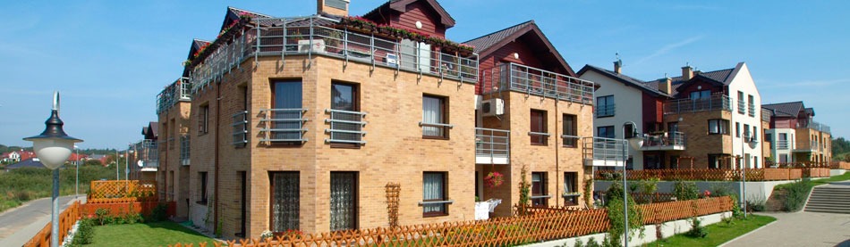 Kompleks budynków mieszkalnych z cegły klinkierowej, balkony z metalowymi balustradami, kwiaty w donicach na balkonach i przy ogrodzeniu, zadbany trawnik i oświetlenie uliczne.