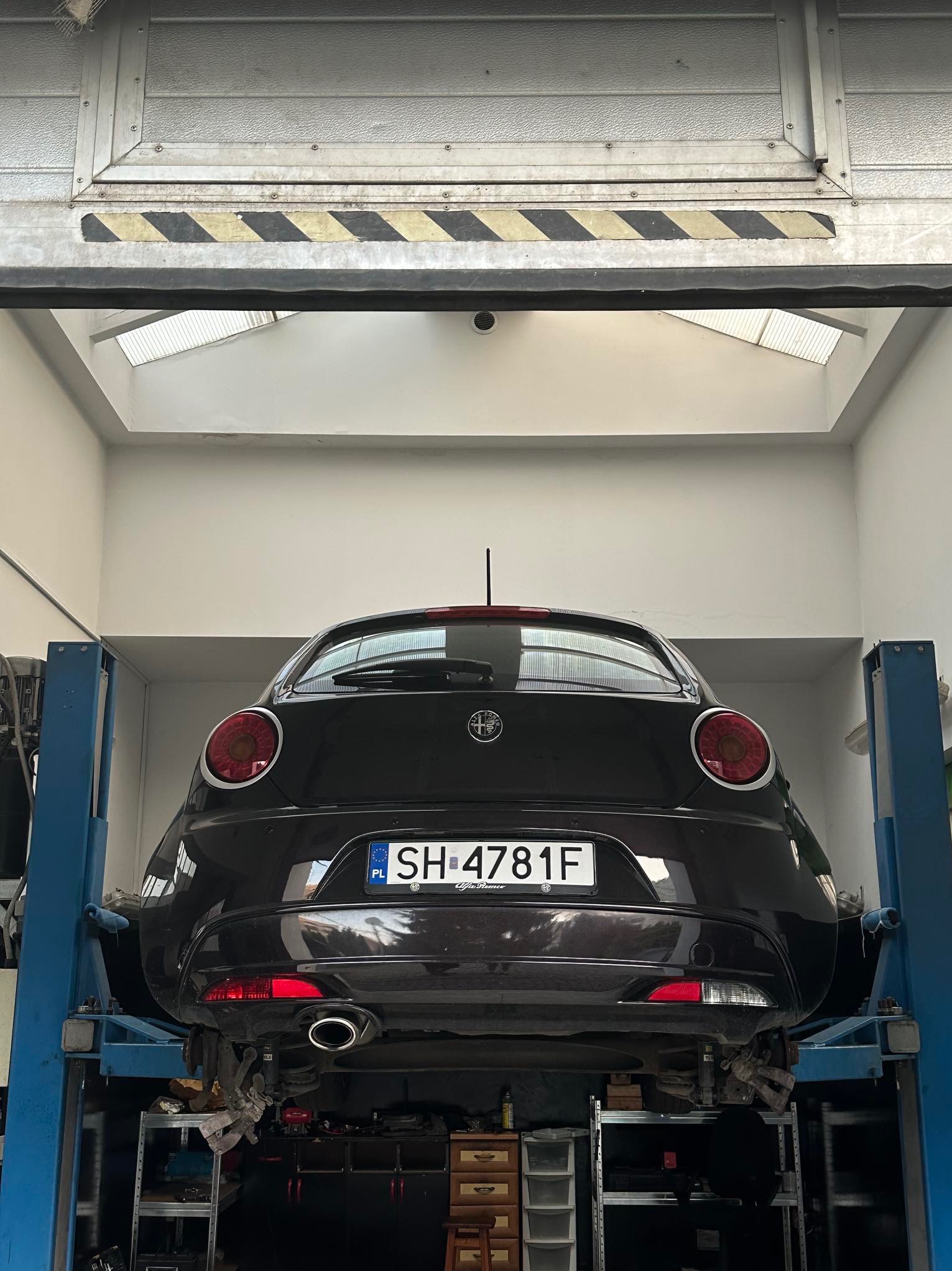 Czarny samochód Alfa Romeo na podnośniku w warsztacie, widok z tyłu, z widocznymi elementami zawieszenia i tablicą rejestracyjną SH-4781F, oświetlenie górne z okna dachowego.