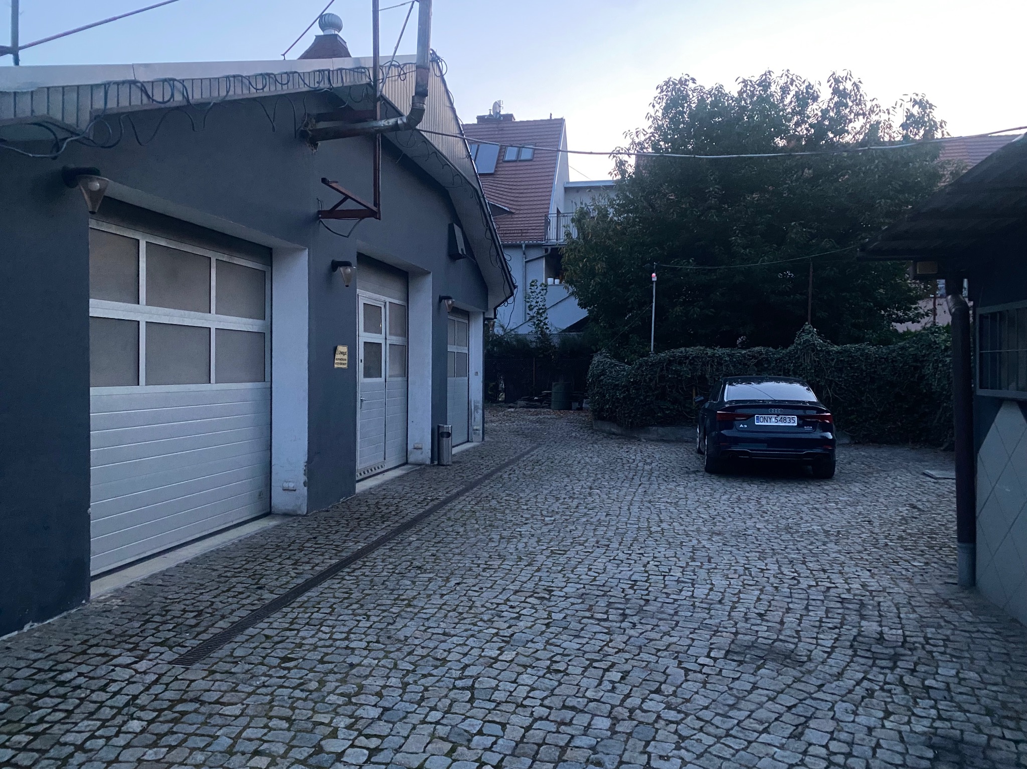 Szary budynek warsztatu samochodowego z trzema bramami garażowymi, wyłożony brukowaną kostką podjazd, zaparkowany ciemny samochód osobowy w tle, widoczne detale architektoniczne i oświetlenie...