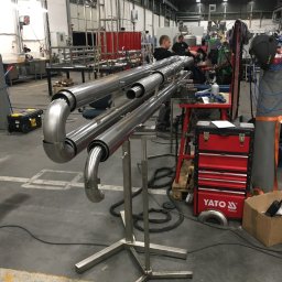 K.K.W. Krzysztof Kotlarz - Konstrukcja z rur ze stali nierdzewnej na metalowym stojaku w hali produkcyjnej, widoczni pracownicy w tle, narzędzia i skrzynka narzędziowa Yato.