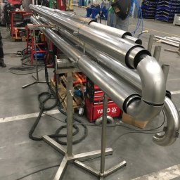 K.K.W. Krzysztof Kotlarz - Konstrukcja z polerowanych rur nierdzewnych na metalowym stojaku, z widocznymi spawami i zagięciami, w tle warsztat z narzędziami i osłoną spawalniczą.