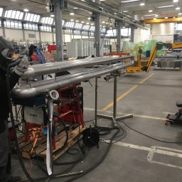 K.K.W. Krzysztof Kotlarz - Pracownik w masce spawalniczej 3M 9100 Speedglas 9100XXi wykonuje spoinę na nierdzewnych rurach w hali produkcyjnej, widoczny wózek spawalniczy EWM Pico 300 Cel puls.