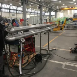 Pracownik w masce spawalniczej 3M 9100 Speedglas 9100XXi wykonuje spoinę na nierdzewnych rurach w hali produkcyjnej, widoczny wózek spawalniczy EWM Pico 300 Cel puls.