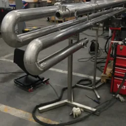 Konstrukcja z rur nierdzewnych na metalowym stelażu w warsztacie, widoczne ślady obr&oacute;bki i spawania, narzędzia i akcesoria w tle.