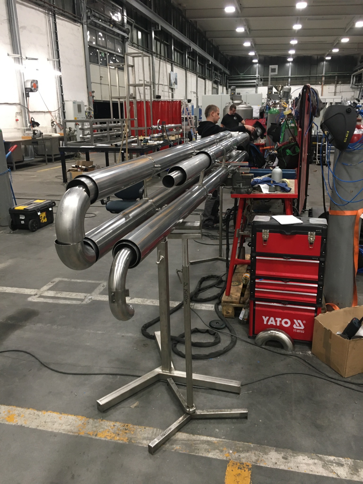 Konstrukcja z rur ze stali nierdzewnej na metalowym stojaku w hali produkcyjnej, widoczni pracownicy w tle, narzędzia i skrzynka narzędziowa Yato.